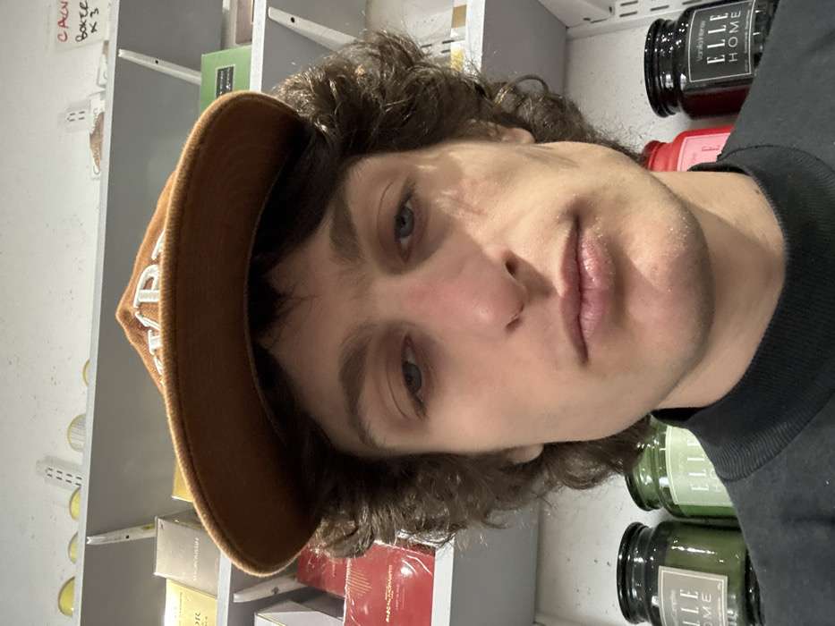 A close-up, front-facing photograph depicts a young adult Caucasian male, approximately 20-30 years of age, with dark curly hair and blue eyes, wearing a dark-colored crewneck shirt and a brown baseball cap with white lettering worn backward. His facial expression is neutral. The subject is situated indoors within what appears to be a retail or storage environment in Joinville-le-Pont, France. Behind the subject, white shelving units are stocked with various commercial products. On the left upper shelves, multiple packaged boxes are visible, including items labeled "MAUBOUSSIN" and "PASCAL MONACO JARDIN RED." On the right upper shelf, a handwritten label on a piece of paper reads "CALN BOXER K3." On the lower shelves, behind the subject's shoulders, several glass jars are present: two green-tinted jars are visible to the left, one clearly marked "ELLE HOME Coconut Candle," and a dark-colored jar to the right is labeled "ELLE HOME Vanilla Intense." The scene is illuminated by uniform artificial lighting.
