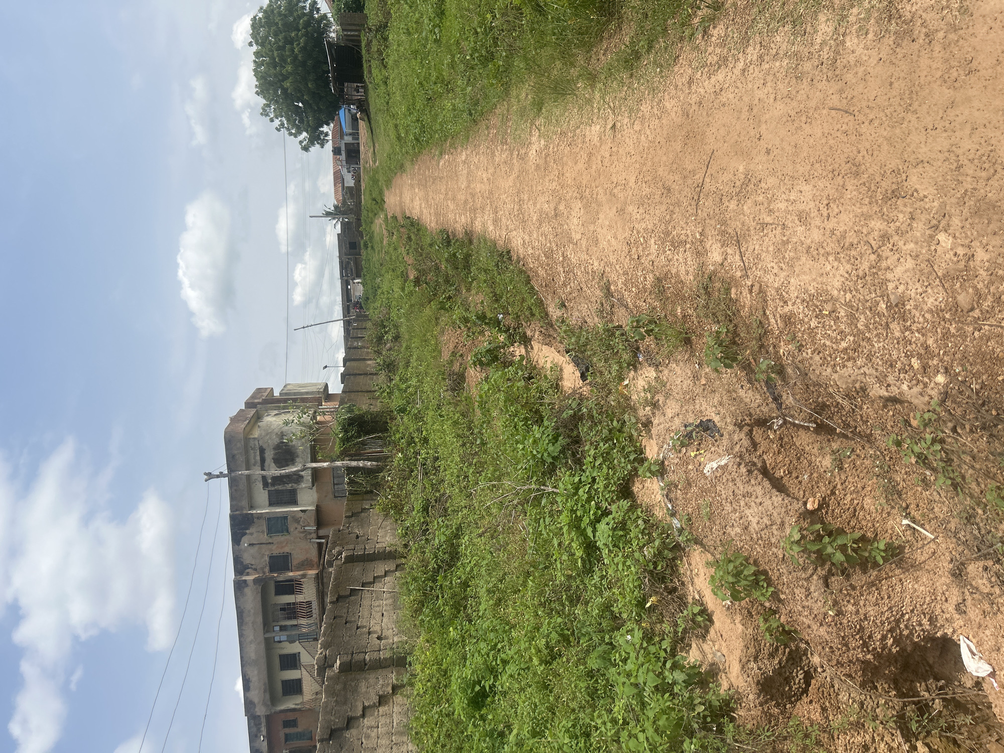 The image depicts an outdoor scene in Ogbomosho, Nigeria, featuring a dirt path leading through overgrown vegetation. On the left, a dilapidated, multi-story building with weathered walls and numerous windows stands behind a low, rough-hewn stone wall. The building appears abandoned or in a state of disrepair, with peeling paint and visible structural damage. The path itself is a light brown dirt track, flanked by tall green grass and weeds. To the right of the path, more greenery continues, with a large tree visible in the distance. In the background, other structures, some appearing to be in various stages of construction or disrepair, can be seen, along with power lines stretching across the sky. The sky is partly cloudy with a mix of white clouds and blue sky. The overall environment suggests a rural or semi-urban setting with evidence of both natural growth and human habitation, though the condition of the buildings indicates a lack of recent maintenance.