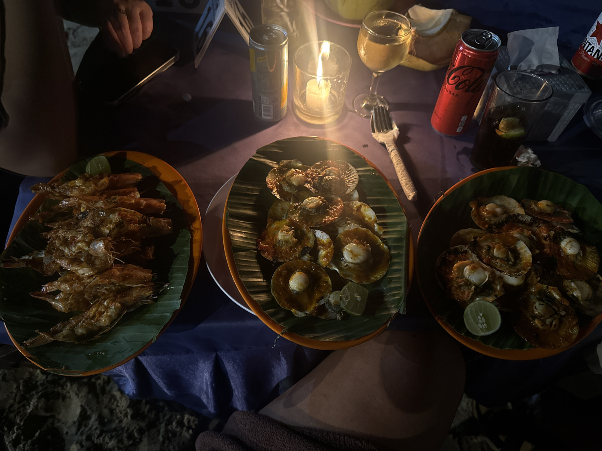 The scene is a dimly lit outdoor dining area, likely on a beach in Kuta Selatan, Indonesia. A table covered with a blue tablecloth holds three orange-rimmed plates, each featuring a different seafood dish arranged on a green banana leaf. The leftmost plate contains what appears to be grilled shrimp. The middle plate displays scallops with a glossy sauce. The rightmost plate also contains scallops, but with a different preparation. A lime wedge is visible on two of the plates. Beverages, including a wine glass filled with a light-colored liquid, a can of Coca-Cola Zero Sugar, and a glass with a dark liquid and lime slice, are also on the table. A lit candle in a glass holder and a can of Bintang beer add to the ambiance. Parts of two people are visible: a hand holding a smartphone and a portion of a leg. The background shows hints of sand.