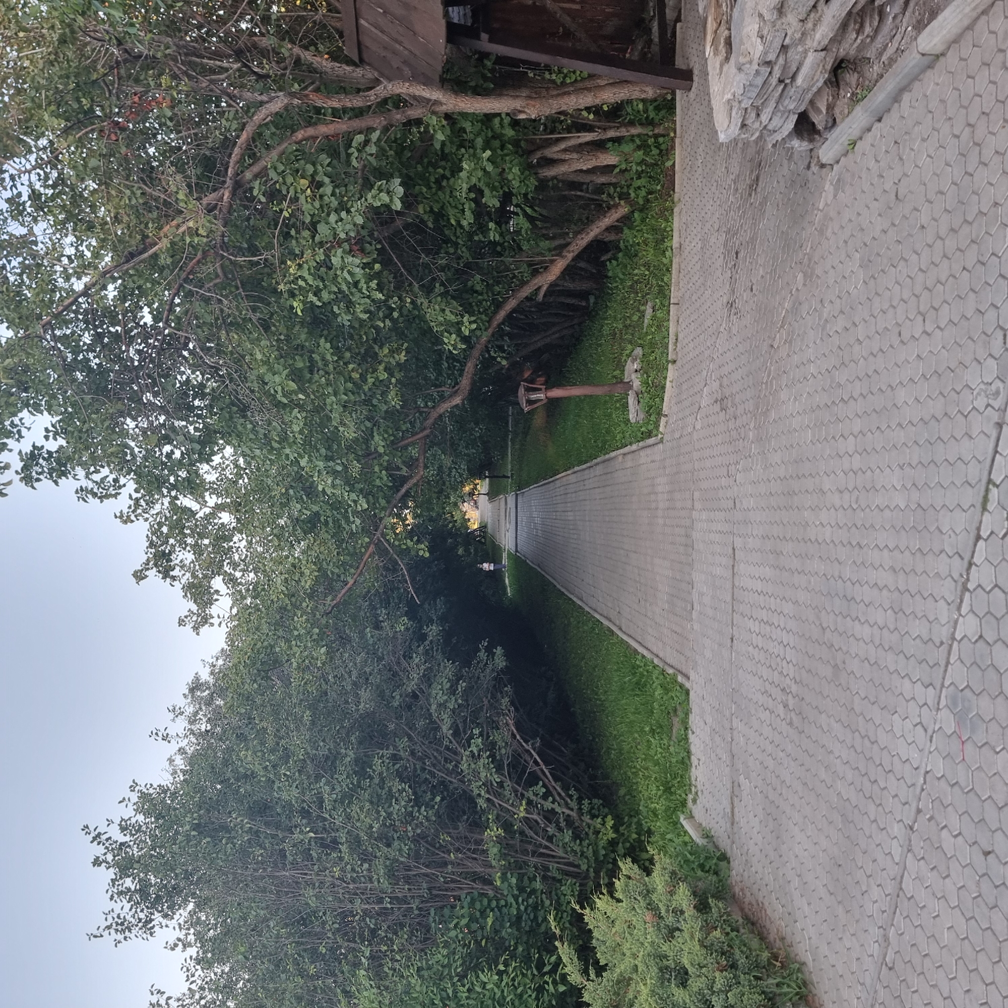 An eye-level, medium shot shows a paved walkway in what appears to be a park or garden area, possibly located in the city of Aktobe, Kazakhstan. The walkway is made of interlocking hexagonal concrete pavers and extends into the distance, disappearing under a canopy of trees. On either side of the walkway, there is lush green grass and dense foliage. In the foreground, on the right side of the image, is a low stone wall made of stacked, irregularly shaped stones. A dark wooden structure, possibly a gazebo or part of a building, is partially visible behind the stone wall and the trees. A rustic-looking lantern with a wooden post stands near the stone wall, positioned on the grass. In the distance, further down the walkway, a lone person wearing a light-colored shirt and dark pants is visible, standing to the left of the path. The sky above is a pale, overcast gray.
