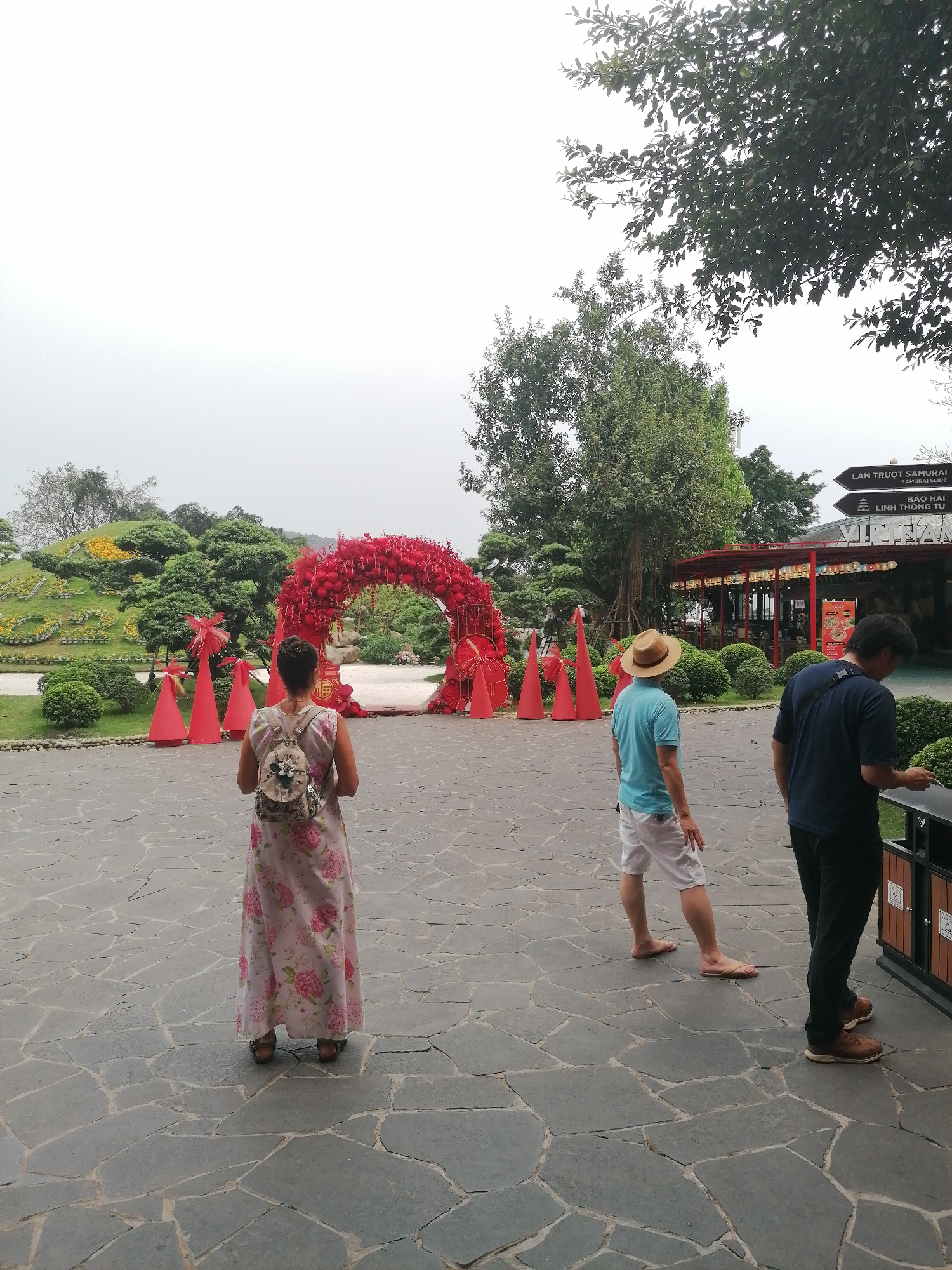 A wide shot captures a courtyard scene at the Lan Truot Samurai Samurai Slide destination in Hong Gai Ward, Vietnam. A woman in a floral print dress and backpack faces a red-decorated archway with conical red structures flanking it. To her right, a man in a blue shirt, white shorts, and a straw hat walks away from the archway. Another man, in a navy blue shirt and dark pants, stands near a trash receptacle. The courtyard is paved with irregularly shaped gray stones. Lush green landscaping includes bushes and trees, one with a sign indicating directions to various attractions, including Lan Truot Samurai and Bao Hai Linh Thong Tu. A building with a red roofline and open-air structure is in the background. The sky is overcast.