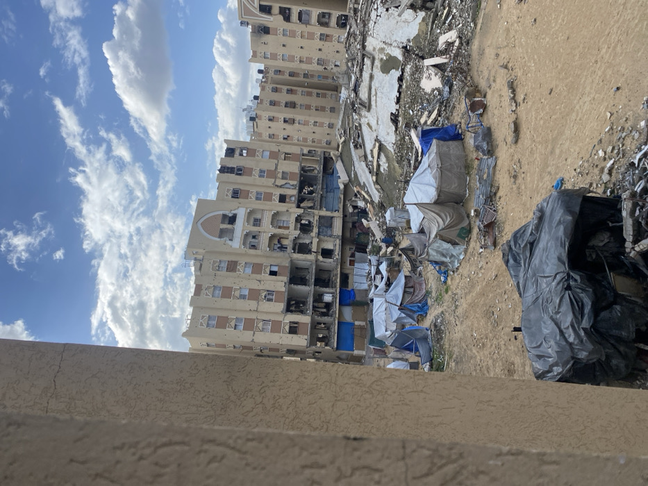 This image, taken in Khan Yunis, Palestinian Territory, captures a scene of severe urban destruction under a bright, partially cloudy sky.
In the background, several multi-story buildings stand heavily damaged. Their facades, primarily light-colored with decorative elements, are extensively scarred. Numerous windows are shattered, missing, or blackened by fire, and large sections of the buildings show structural compromise, with interior spaces exposed. Pockmarks, possibly from bullets or shrapnel, are visible across their exteriors. On the roofs of these buildings, water tanks are visible, some appearing intact while others may be damaged.
The ground in the mid-ground and foreground is completely covered in a vast expanse of rubble, broken concrete, and debris, indicating that other structures have been demolished or collapsed. Amidst this devastation, several makeshift shelters constructed from various tarpaulins—some blue, others grey or off-white—are scattered across the dusty, uneven terrain. These rudimentary shelters suggest temporary living arrangements in the destroyed area. A large black tarp or plastic sheet covers an unknown object in the lower right foreground.
The time of day appears to be daytime, with natural light illuminating the scene and casting soft shadows. The sky is bright blue with scattered white clouds. No people are visible in the image. The overall impression is one of a post-conflict or disaster-stricken landscape, marked by widespread destruction and the presence of temporary dwellings. No visible text is discernible in the image.