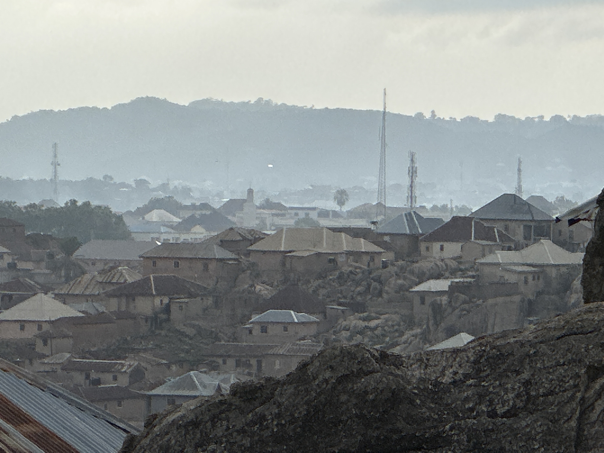 The image shows an aerial view of a densely built neighborhood in Jos, Nigeria, characterized by closely packed, single-story houses with varying roof materials. The houses appear perched on rocky terrain, with visible rock formations in the foreground. In the distance, a hazy outline of hills is visible, punctuated by several communication towers. The sky is overcast, contributing to the muted color palette and hazy atmosphere. The overall impression is one of a densely populated area built within a rugged, hilly landscape.