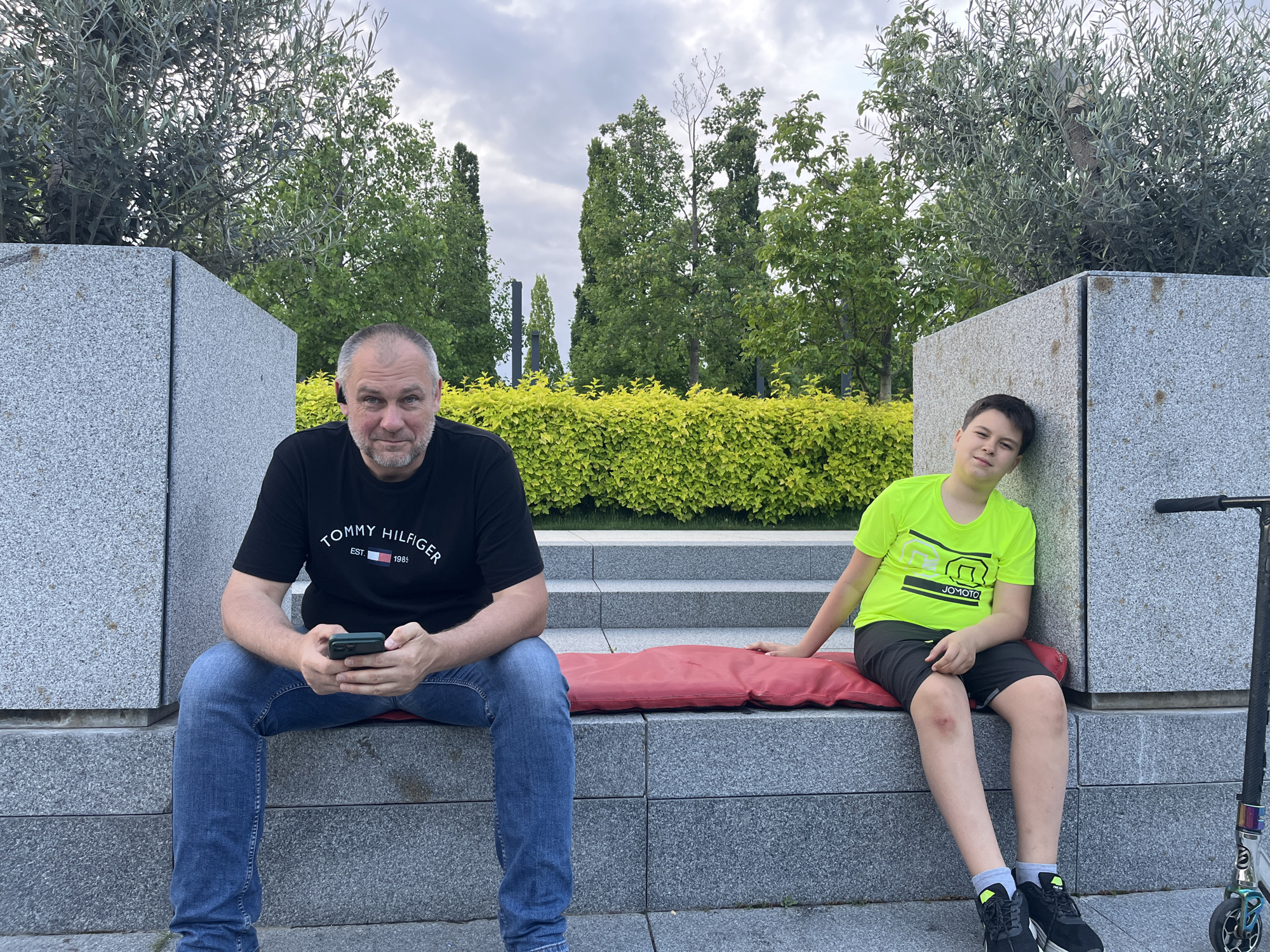 A middle-aged man sits on a stone bench in a park, looking at the camera while holding a smartphone. He wears a black Tommy Hilfiger t-shirt and blue jeans. A young boy in a neon green t-shirt and black shorts leans against a stone planter to the man's right. A red cushion lies between them on the bench. A silver scooter stands near the right edge of the frame. The background features green trees and shrubbery under an overcast sky. The scene is located in Krasnodar, Russia.