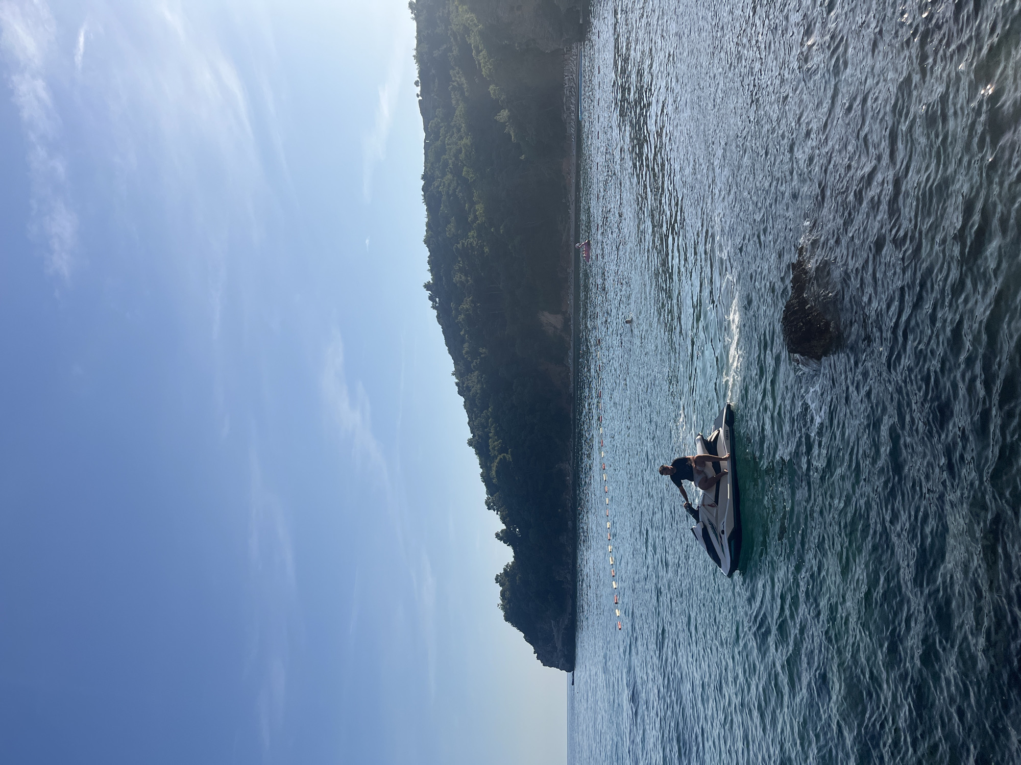 The image displays a bright, sunny coastal scene in Budva, Montenegro, featuring a person on a jet ski in the foreground against a backdrop of a lush, forested mountain and clear blue waters.
The sky, occupying the upper left portion of the frame, is a vibrant blue with faint, wispy clouds. A large, verdant mountain or cliff face, densely covered in trees and foliage, stretches across the mid-ground, rising steeply from the water. The sunlight strongly illuminates the scene, casting soft shadows.
In the foreground and mid-ground, the water is a deep blue-green, showing distinct ripples and reflections from the sun. A white and black jet ski with a single person standing on it is positioned towards the right side of the frame, appearing ready for activity. A line of white and orange buoys extends across the water, likely indicating a swimming or activity zone. A dark rock is partially submerged in the water, closer to the center. In the distant water near the base of the mountain, small, faint figures suggest other people swimming or engaging in water recreation.
The scene portrays a leisurely and active day, with excellent weather conditions for water sports and enjoyment of the natural coastal environment. The time of day appears to be mid-day, given the bright, direct sunlight. No discernible text is visible in the image.