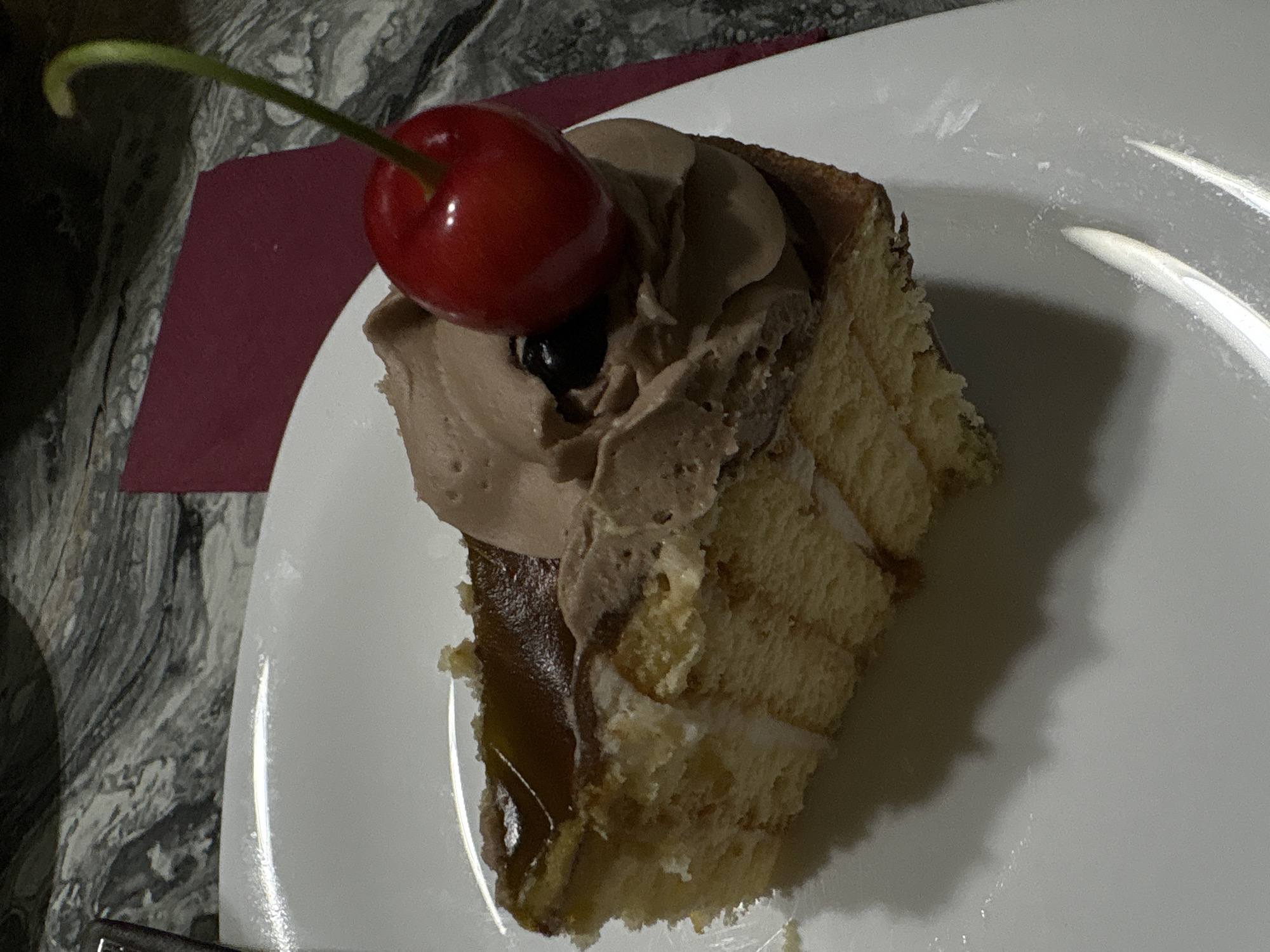 A slice of chocolate cake with whipped cream and a cherry on top sits on a white plate. The cake has layers of sponge cake and cream, with a glossy chocolate topping. A single red cherry with a green stem rests on the whipped cream. The plate is on a dark, marbled surface. A maroon napkin is partially visible to the right of the plate. The image is well-lit, highlighting the textures and colors of the dessert. The location is specified as the city of Dalgopol, Bulgaria.