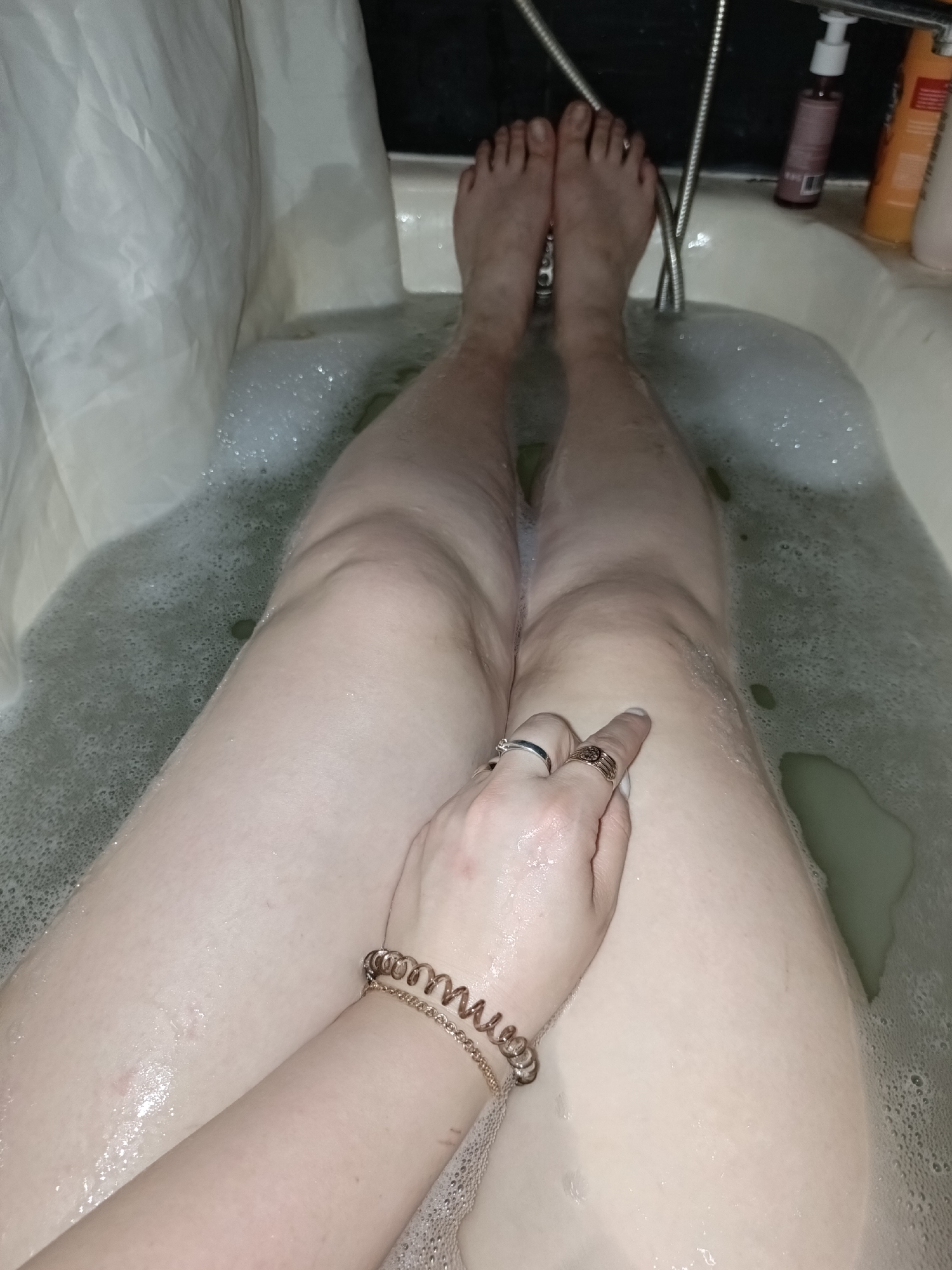 The image captures a first-person perspective of a person's legs and feet submerged in a bubble bath. The setting is a bathroom, with the lower half of the person's body visible from the waist down in a white bathtub filled with bubbly, slightly murky water. The person's feet are visible at the far end of the tub, resting near a shower hose. On the right side of the image, several bottles of toiletries (shampoo, soap, etc.) are lined up on the tub's edge.
Closer to the viewer, a hand is resting on the right thigh. The hand wears two rings: one silver-colored band on the ring finger and a gold-colored, intricately designed ring on the middle finger. Two bracelets are visible on the wrist: one appears to be a coiled, spring-like design in rose gold or copper tone, and another is a delicate gold chain. A white shower curtain is drawn on the left side of the tub. The lighting suggests an indoor setting, likely artificial light. No visible text is discernible on any objects.