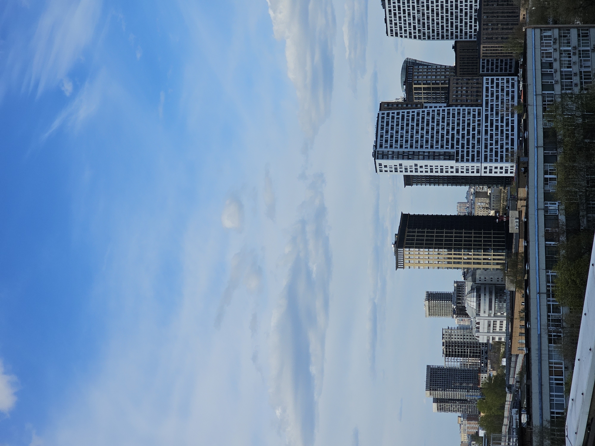 The image presents a panoramic view of a modern cityscape in Moscow, Russia, dominated by numerous tall buildings against a bright blue sky. The perspective suggests the photo was taken from an elevated position, possibly a bridge or an upper floor, with a white railing or structure visible in the lower right foreground.
The right side of the frame is densely packed with various urban structures. Many buildings are contemporary, featuring diverse architectural styles and materials. Several prominent high-rises exhibit grid-like patterns of windows, some with white facades and dark windows, while others appear darker, possibly constructed from glass or dark stone. One distinctive building has a rounded or domed roof visible among the taller structures. Further into the background, more buildings of varying heights and designs stretch across the urban landscape. Patches of green foliage, likely trees, are visible between and in front of some buildings, providing a touch of nature amidst the concrete.
The left side and upper portion of the image are filled with a clear, vibrant blue sky, dotted with scattered white and wispy clouds. The lighting indicates it is daytime, likely morning or afternoon, given the clarity and natural illumination of the scene. No specific human activity is discernable, nor are any individuals visible. There is no visible text or identifiable landmarks beyond the general modern architecture of a large city. The overall impression is one of expansive urban development under a serene sky.