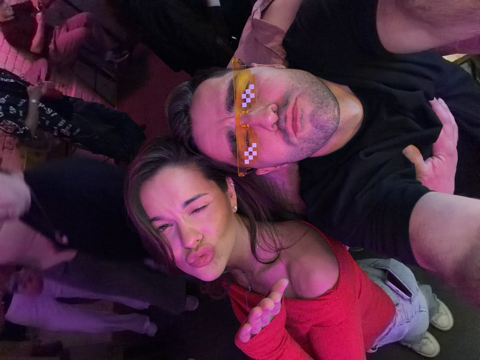 A tilted selfie captures a man and a woman in a dimly lit indoor setting, strongly suggested to be a nightclub or party in Krasnoyarsk, Russia, due to the prominent pink and purple ambient lighting.
The man, positioned on the right side of the frame, wears a black long-sleeved shirt. He sports short facial hair and distinctive yellow, pixelated novelty sunglasses, resembling "deal with it" meme glasses. His expression is mostly neutral with a slight pout. His left arm is visible, seemingly around the woman or holding her close.
The woman, in the foreground on the left, has dark hair and is wearing a bright red long-sleeved top, possibly off-shoulder, paired with light-wash jeans and white sneakers. She is actively posing for the camera, making a kissing face with pouted lips and winking with her right eye. Her right hand is raised towards the camera, fingers slightly curled, adding to her playful demeanor. She wears earrings.
The background is dark and blurred, revealing faint shapes of other people and elements of the venue, all bathed in the vibrant pink and purple glow. On a dark garment in the background, partially legible white text includes "p-91" and "TICA". The image conveys a lively, late-night social atmosphere.