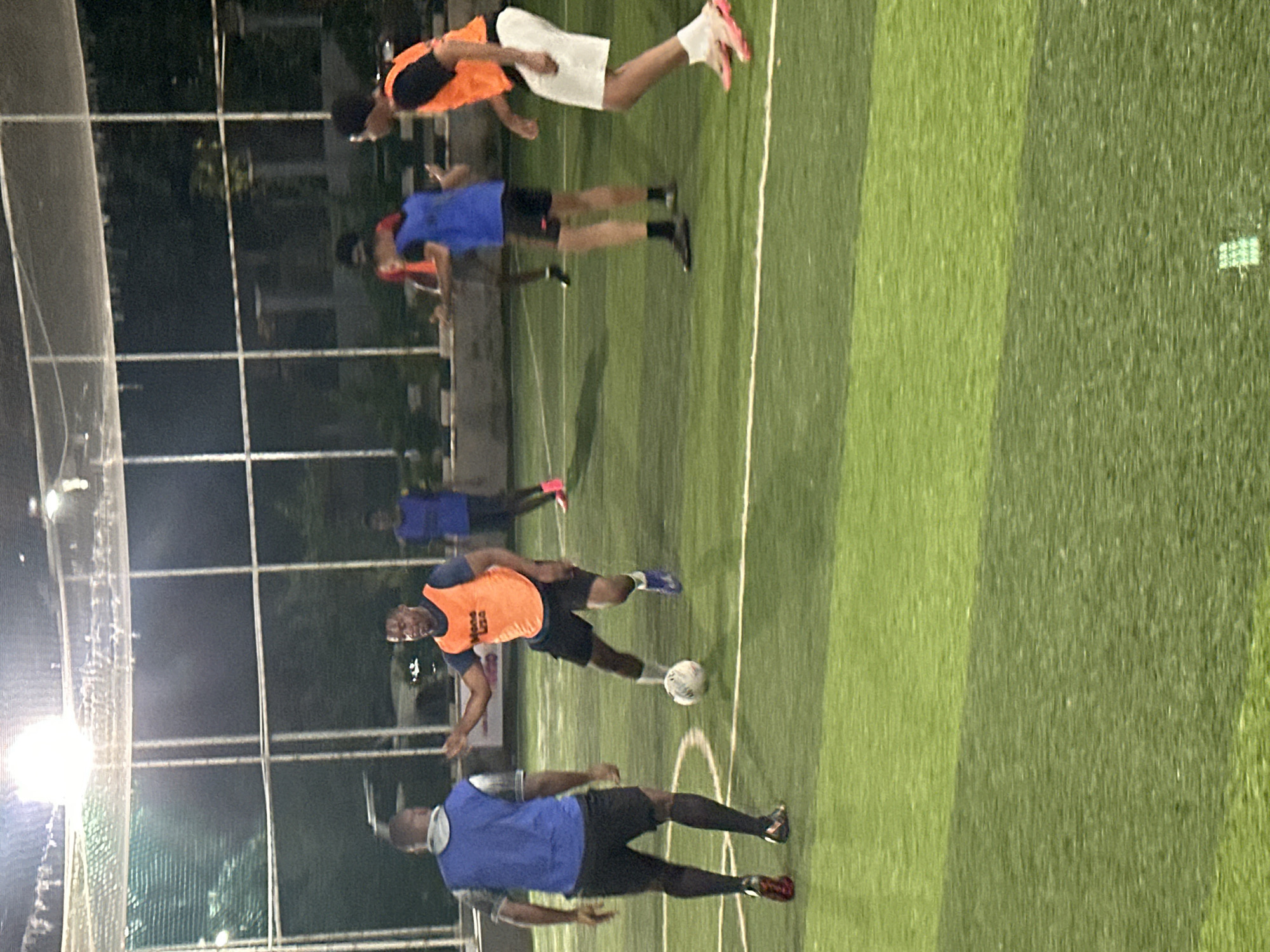 A group of men are playing soccer on an artificial turf field at night. The field is enclosed by a fence and lit by overhead lights. In the foreground, a man wearing a blue vest and black shorts is facing away from the camera, while another man in an orange vest and black shorts is in possession of the ball, which is on the ground between them. To the right, a man in an orange vest and white shorts is running. In the background, several other men in various colored vests are also visible on the field. The scene appears to be at the Abuja National Stadium, which is a sporting complex located in the capital city of Nigeria.