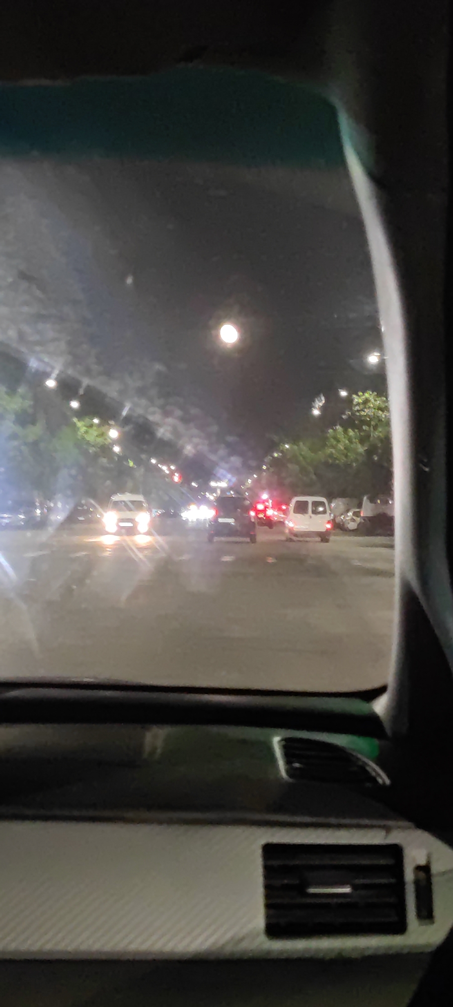 This nighttime image is captured from inside a vehicle, looking out through the windshield onto an urban street in Yerevan, Armenia. The scene depicts a road illuminated by numerous artificial lights and a striking, bright circular light high in the dark sky, resembling a full moon or a powerful street lamp.
In the foreground, parts of the car's interior are visible. The dashboard features a distinct white, patterned surface, possibly a carbon fiber wrap, on the left side, and a dark air vent on the right. The car's interior pillars frame the view, narrowing the perspective.
The road ahead appears wet and highly reflective, suggesting recent rain. Headlights and taillights from various vehicles create bright streaks and reflections on the dark asphalt. A white van or minibus is clearly visible in the middle distance, its red taillights glowing. Another car directly in front of the van displays bright headlights. Streetlights are scattered along the road, casting light on the green trees that line both sides of the street. The overall impression is of a moderately busy city street at night, with a damp atmosphere. No discernible text is visible in the image.