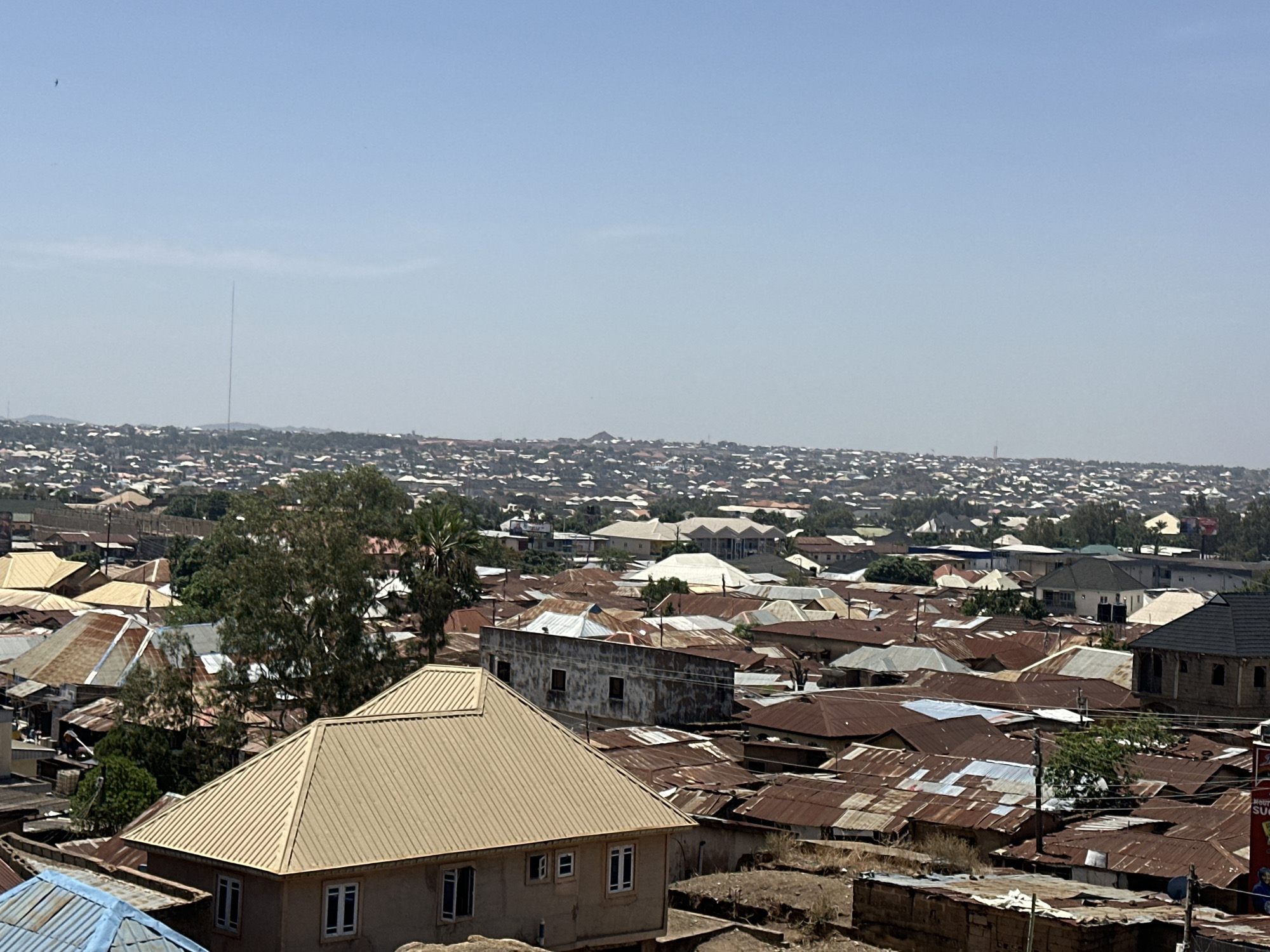 The image depicts a panoramic view of Jos, Nigeria, showcasing a dense urban environment. A multitude of buildings with corrugated iron roofs dominate the landscape, ranging from rusty brown to silver hues. A new tan-colored building with a gabled roof stands out in the foreground. Patches of greenery are visible, interspersed amongst the rooftops. In the distance, a tall communication tower pierces the horizon, situated against a hazy sky.