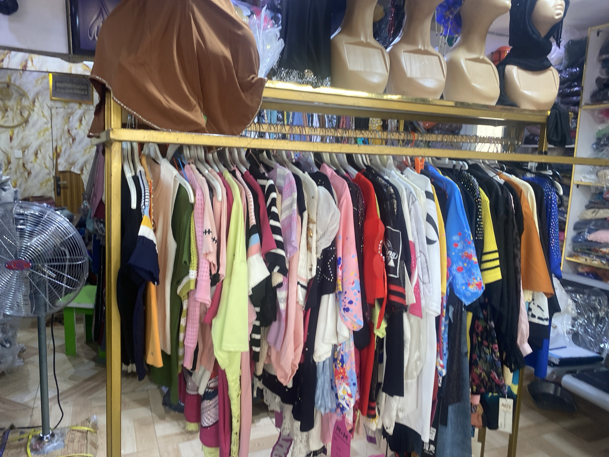 The image captures the interior of a retail clothing store, likely located in Lagos, Nigeria. A gold-colored clothing rack dominates the foreground, densely packed with a variety of garments hanging on individual hangers. These include colorful sweaters, shirts, and dresses in shades of pink, green, black, and blue, with some items featuring patterns or embellishments.
Above the clothing rack on a higher shelf, several mannequin heads are displayed. These mannequin heads are light-skinned and bare-headed, but some have headscarves or hijabs.
To the left of the clothing rack stands a silver metal fan. A green stool or table is partially visible behind the fan. The store's interior shows wallpapered walls and other display shelves with additional merchandise in the background. The overall impression is that of a busy, stocked retail environment.