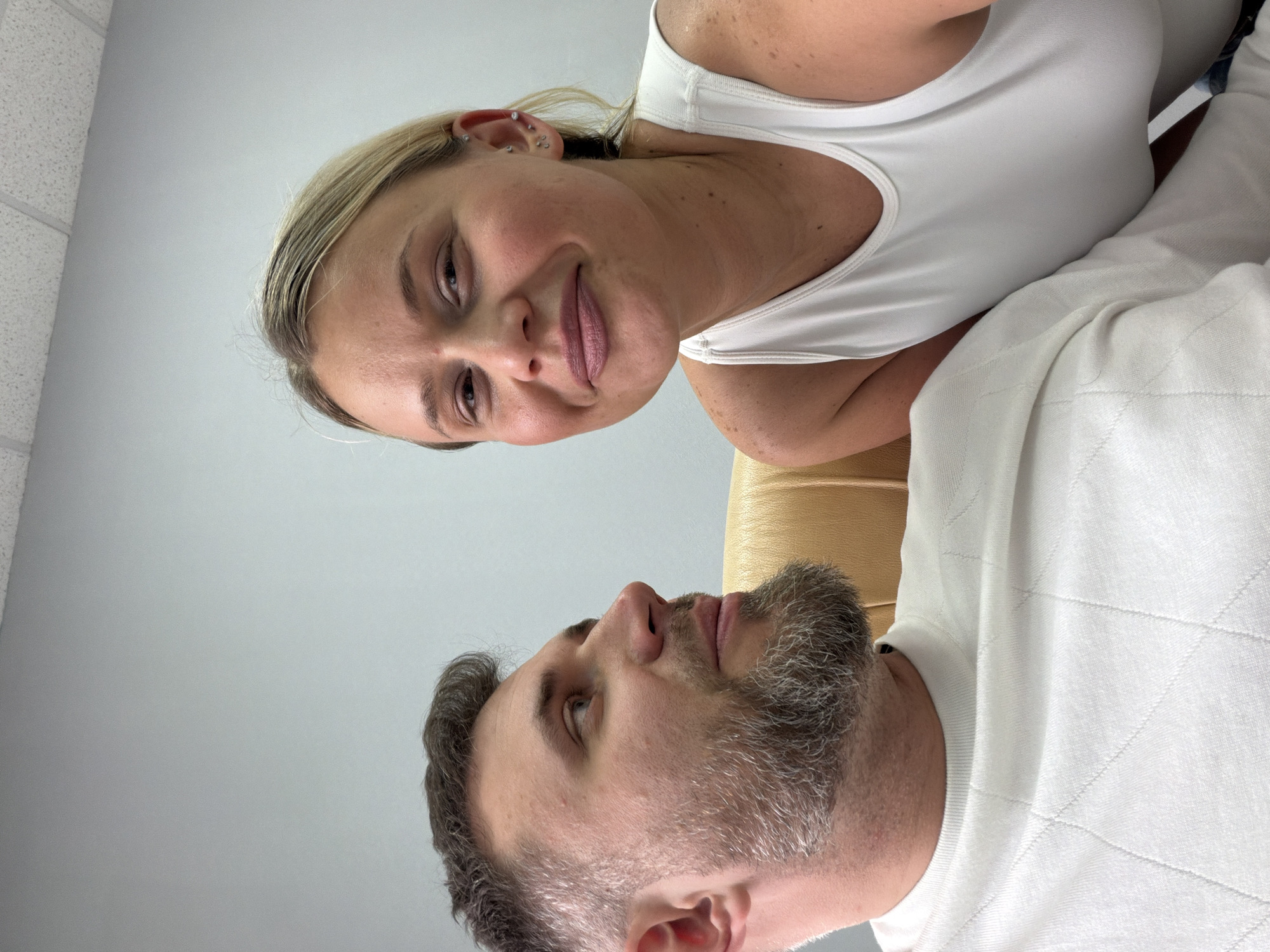 A woman and a man are shown from a low angle, looking down at the camera. The woman has blonde hair and is wearing a white tank top. She has multiple piercings in her left ear. The man has a beard and is wearing a white sweater with a diamond pattern. They are positioned close to each other. The background is a plain light gray wall and a white textured ceiling. The bottom right corner shows a tan-colored surface, possibly part of a chair or couch. There is no indication of the location being Sarapul, Russia.