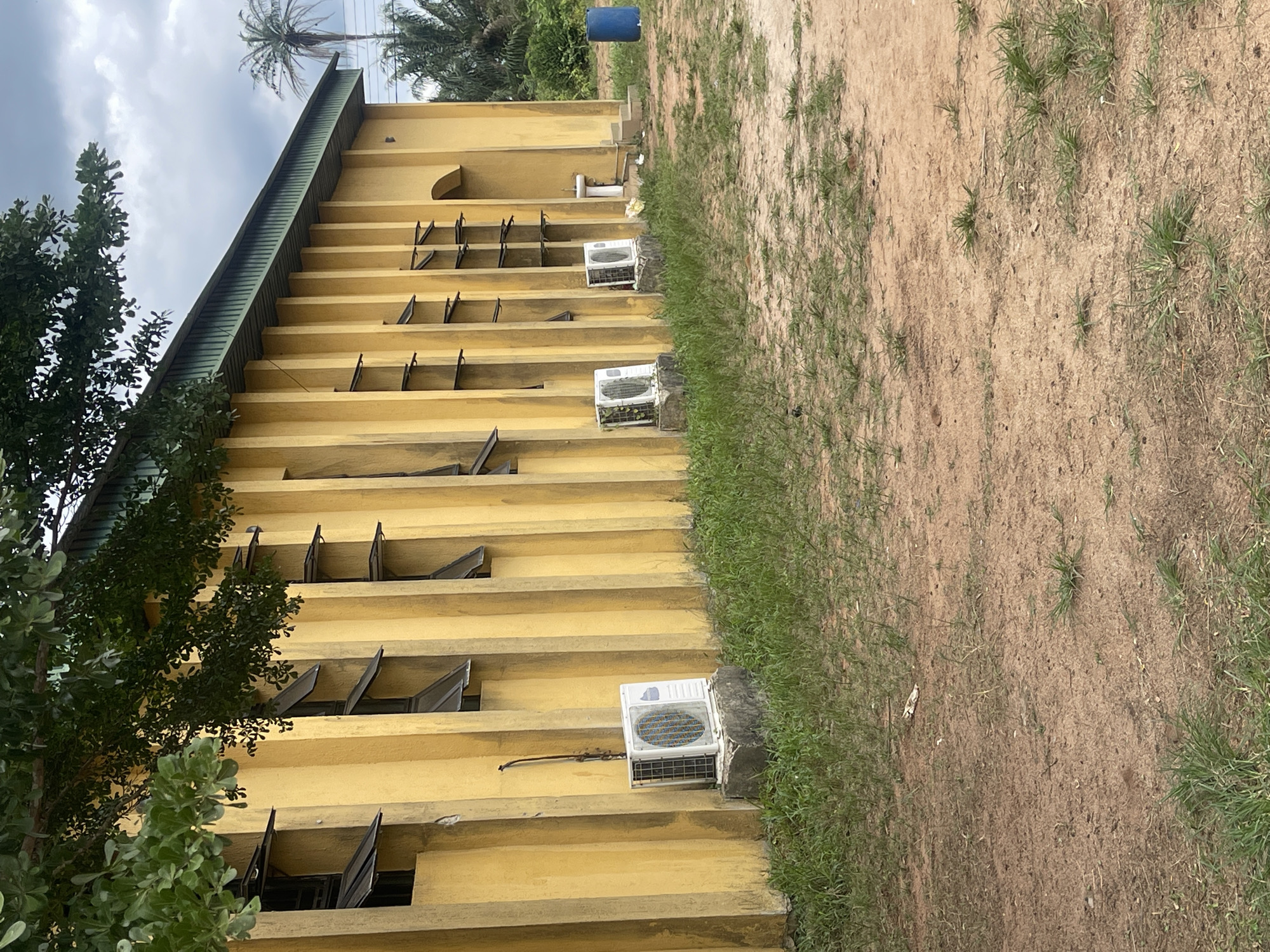 A yellow building with multiple levels and awning-style windows is depicted. Air conditioning units are affixed to the building's exterior. A tree is visible in the foreground, and a grassy area borders the building on the right. The ground is dirt and sparse grass. A palm tree is visible in the background with a blue plastic drum located behind the building. The image appears to be taken in Abraka, Nigeria.