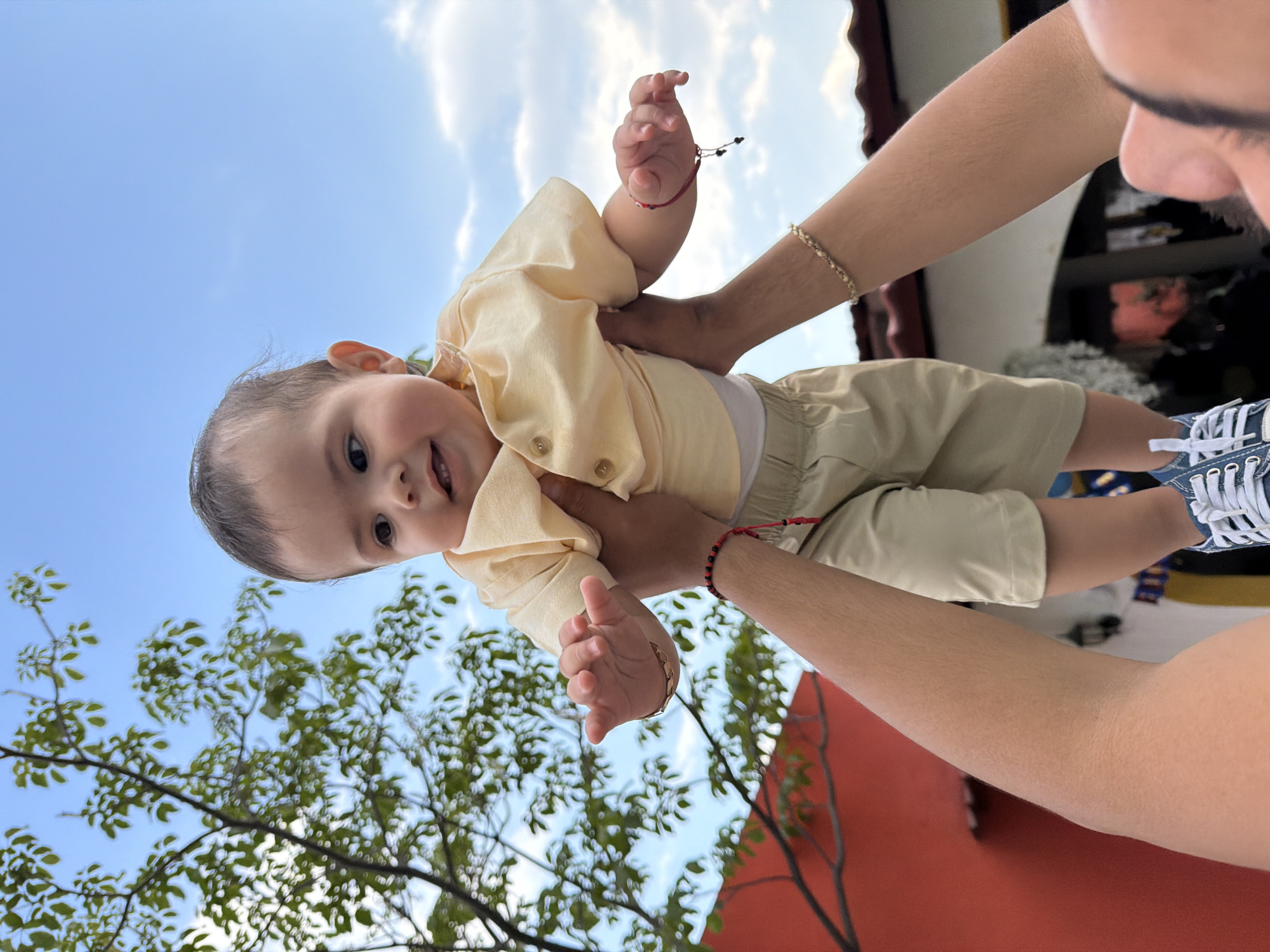 A medium shot captures a baby being held aloft against a sky backdrop, potentially in Cuautla, Mexico. The baby, clad in light yellow attire, is held by adult arms. The adult's face is partially visible, wearing blue sneakers. The background features a blend of blue sky, foliage, and a structure with orange-red walls. A bracelet is visible on the baby's wrist.