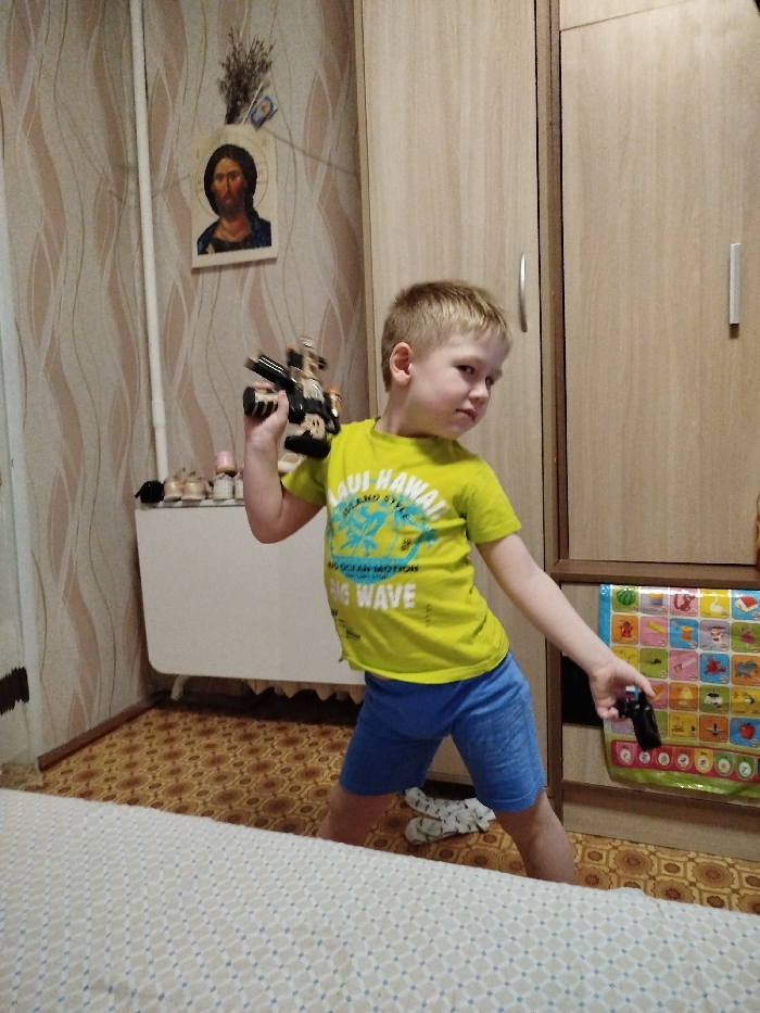 This image, originating from an interior location in the city of Chaikovsky, Russia, depicts a young male child standing in what appears to be a domestic room. The child, with blond hair, is positioned centrally, facing his right with his head turned towards the viewer, adopting a dynamic stance. He wears a lime green short-sleeved t-shirt with a blue "MAUI HAWAII" graphic and blue shorts. In his right hand, he holds a black and gold toy blaster-style weapon, elevated near his shoulder. His left hand, extended downwards, holds a smaller, black toy pistol-like object.
The background consists of a wall covered in beige and white wavy-patterned wallpaper on the left. Mounted on this wall is a rectangular religious icon depicting Jesus Christ, above which a bundle of dried foliage is affixed. Below the icon, a white radiator or low storage unit is visible, with several items, including small shoes, placed on its surface. To the right, a light brown wooden wardrobe unit with two doors and silver vertical handles extends upwards. An educational poster with multiple colorful squares, each containing an image and text, is affixed to the lower part of the right wardrobe door. The floor is covered with a brown linoleum or tile featuring a repetitive circular pattern. A pair of white socks with black patterns lies on the floor near the child's feet. The lower foreground is occupied by a light-colored, patterned sheet, likely covering a bed or large piece of furniture.
