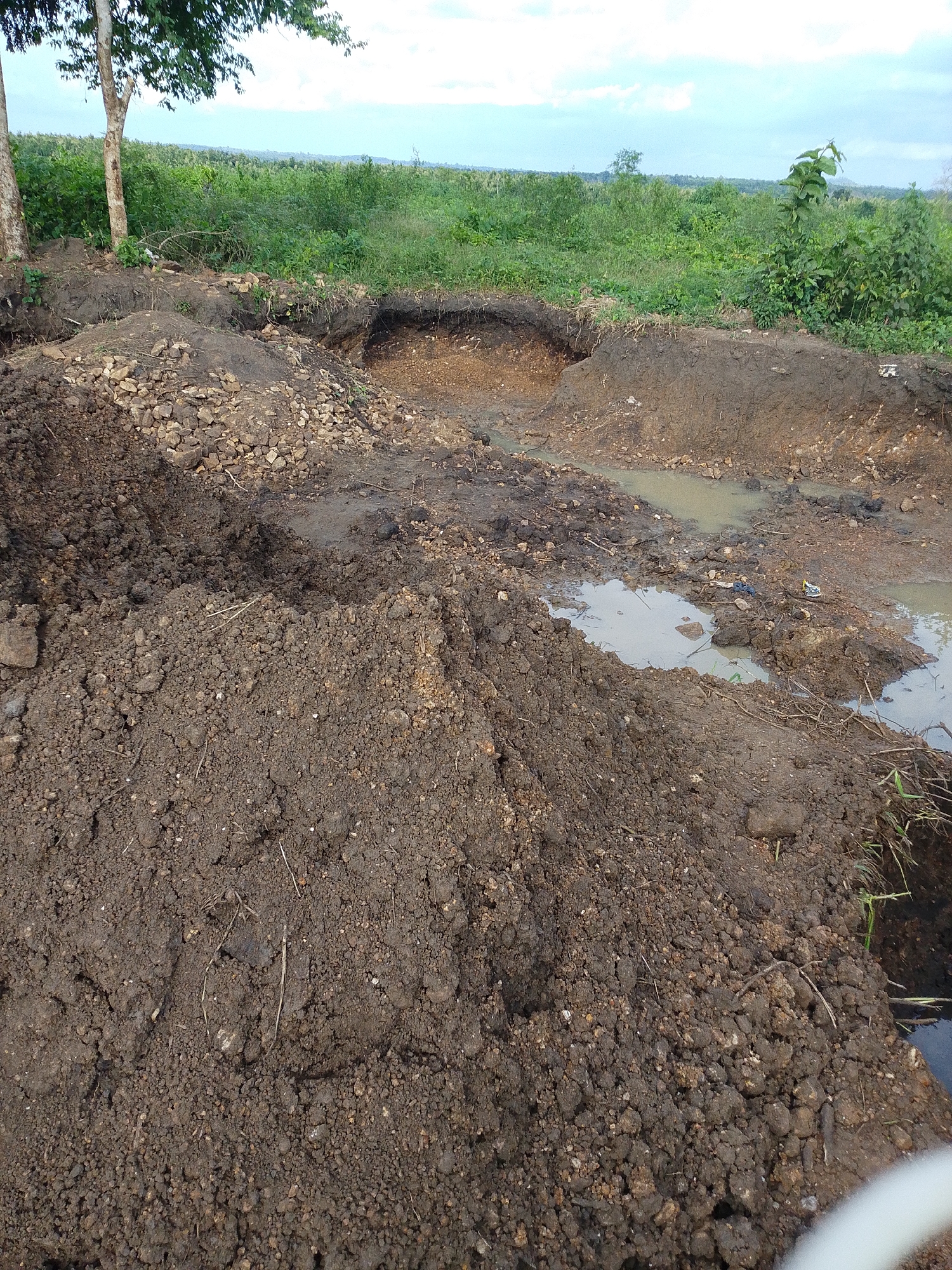 The image shows a disturbed earth landscape with several excavated pits filled with muddy water. Piles of excavated soil and rocks are visible in the foreground and along the edges of the pits. The background consists of dense green vegetation and a cloudy sky. The scene appears to be an outdoor area in Otulu, Nigeria, likely a construction or excavation site. There are no people or animals visible in the image. The overall impression is one of an active or recently active earth-moving operation.