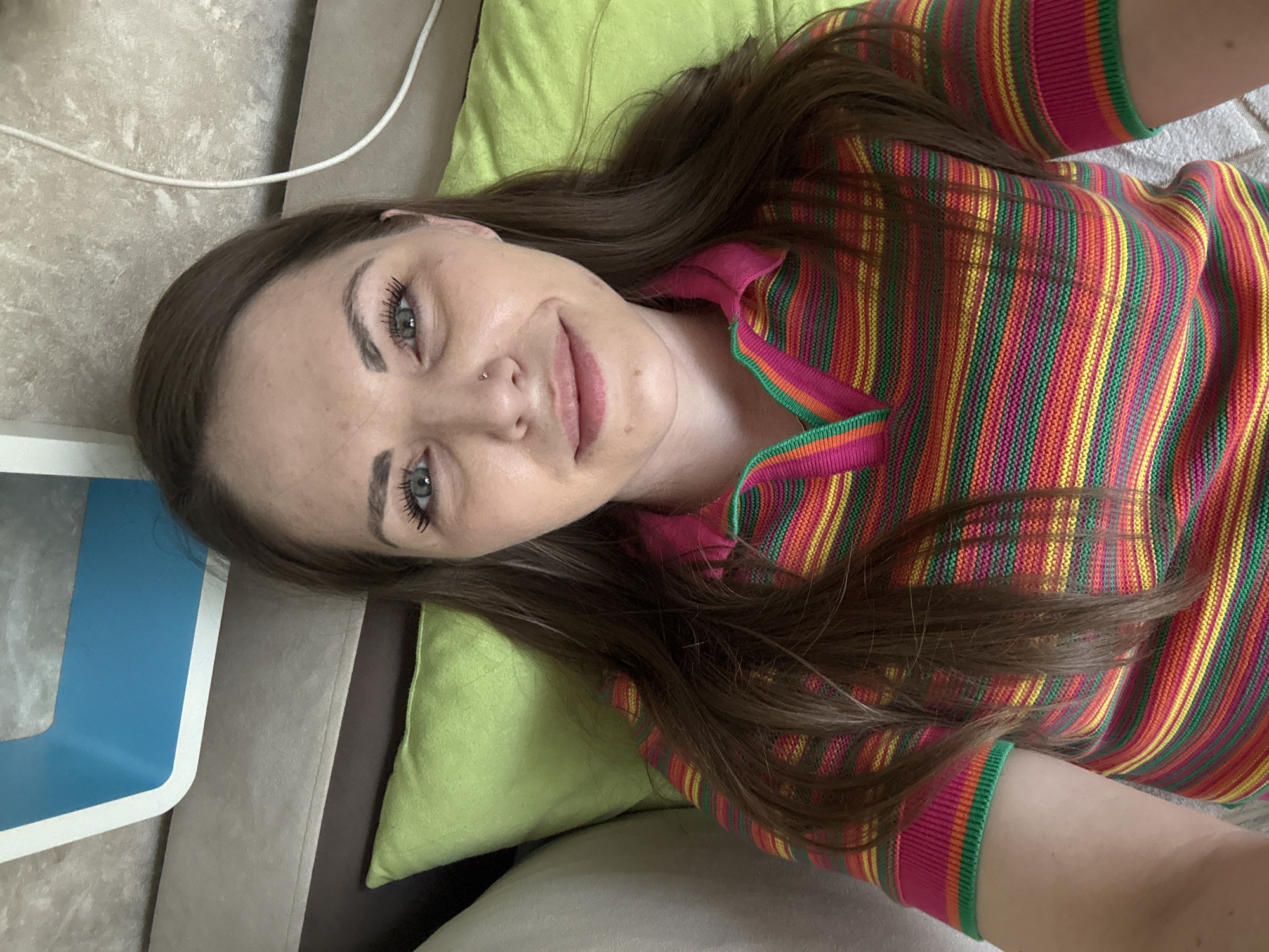 The image shows a woman with long brown hair, lying on a green pillow and a couch. She's wearing a brightly striped pink, orange, yellow, and green shirt. She has light skin, gray eyes, and a nose piercing. The setting appears to be indoors. There is no evidence indicating the location is in Раздолье, Russia.