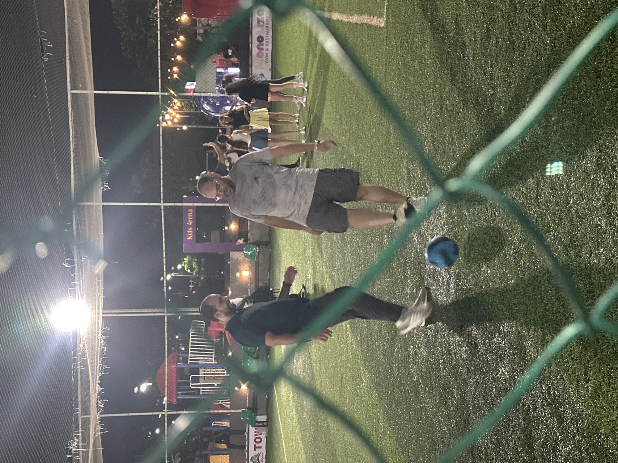 The image shows an outdoor artificial turf soccer field at night in Abuja, Nigeria. Two men are the primary subjects, engaged in playing soccer. One man, wearing glasses and a grey t-shirt with dark shorts, is in the foreground, facing left and appearing to be mid-stride. Another man, in a dark short-sleeved shirt and dark pants, is on the left, also in motion, facing right and looking towards the ball. A blue soccer ball is visible on the ground between them. In the background, several other people, appearing to be a mix of adults and possibly children, are gathered. There is playground equipment with red and blue structures, and string lights illuminating the area. The field is bordered by a green net, part of which is in the foreground, obscuring some of the view. The overall environment suggests a recreational facility.