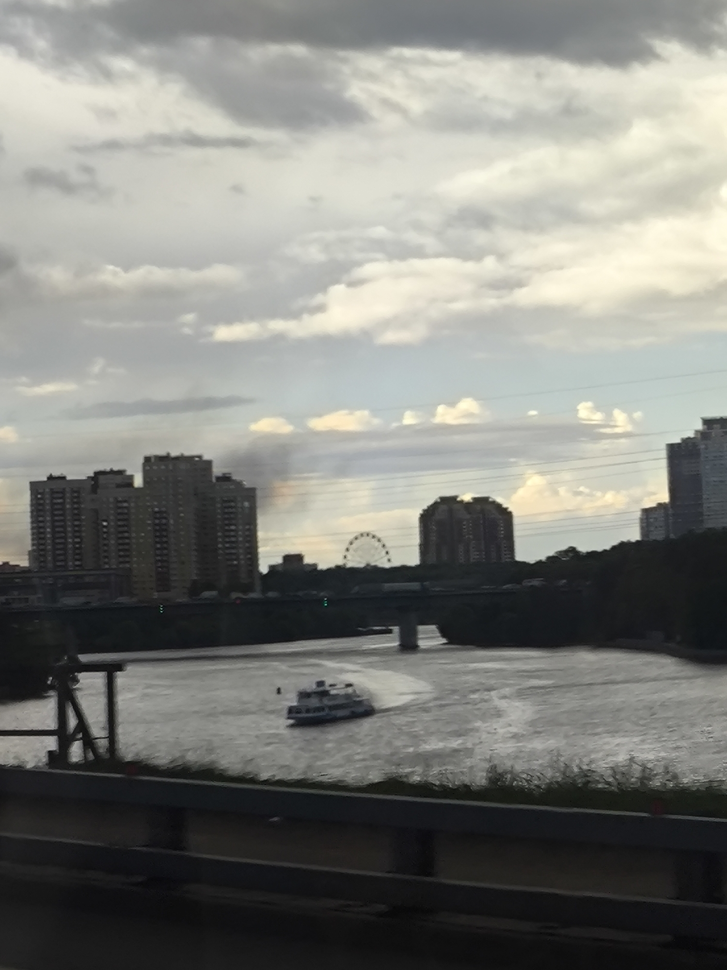 The photograph depicts a river scene with multiple buildings in the background. A passenger ferry boat is navigating the river, creating a small wake. A bridge spans the river, connecting the areas in the foreground and background. The skyline includes high-rise buildings and a Ferris wheel. The sky is filled with clouds. A guardrail is visible in the foreground, suggesting the photograph was taken from a vehicle or elevated platform. It is hard to locate the precise location.