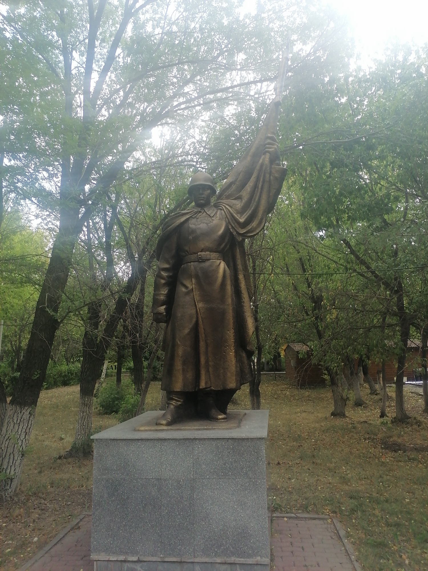 A bronze statue of a soldier stands on a grey granite pedestal in an outdoor setting. The soldier, wearing a helmet and a long coat with a belt, holds a flag aloft with his right hand, which is partially obscured by the draped fabric. The statue is positioned in a park-like environment with numerous trees, some with bare branches and others with green leaves, suggesting either early spring or late autumn. The ground is covered with dry leaves and grass. In the background, there are some wooden structures and more trees. The sunlight is bright and appears to be coming from the upper right of the frame, causing some lens flare. The image appears to be taken in the city of Qarağandı city administration, Kazakhstan, based on the context provided.