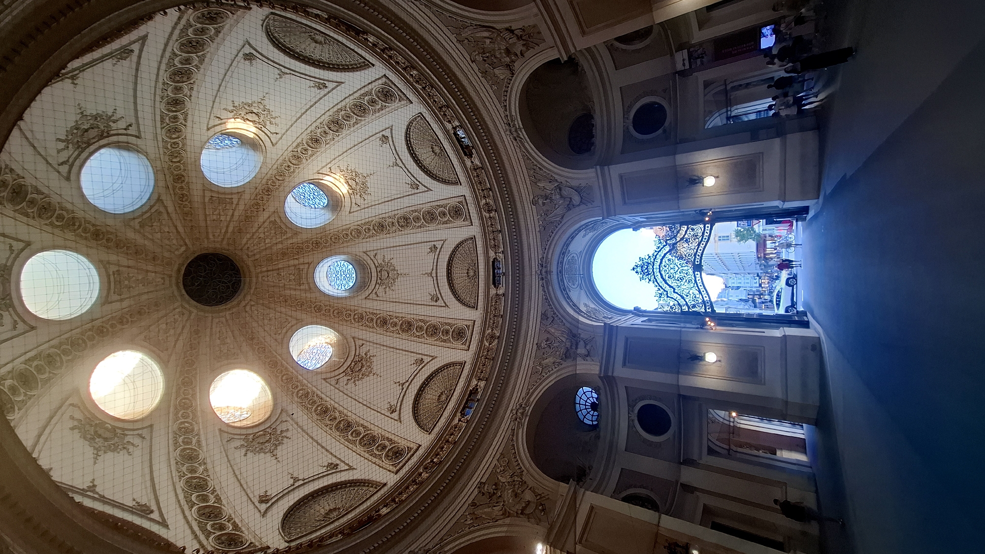 The image shows a wide-angle view looking up at a grand, ornate dome ceiling in a building, and looking out through an archway to a city street in Vienna, Austria. The dome is a pale, creamy color with intricate decorative patterns and a series of round windows that allow natural light to filter in. The windows vary in how brightly they are lit, suggesting different times of day or varying cloud cover. Below the dome, there are large, arched doorways and windows, also decorated with sculpted details.
The archway in the center leads to a street scene. Through the ornate, wrought-iron gate of the archway, buildings with classical architecture and several people are visible. A white SUV is parked on the street, and a few individuals are walking. The street appears to be well-lit, possibly during the early evening or a bright day. On the right side of the archway, several people are gathered, some appearing to be tourists. The floor in the foreground is a dark, reflective surface, possibly polished stone or concrete. The overall impression is one of grandeur and historical significance. The location is likely a public building or a significant architectural landmark in Vienna.