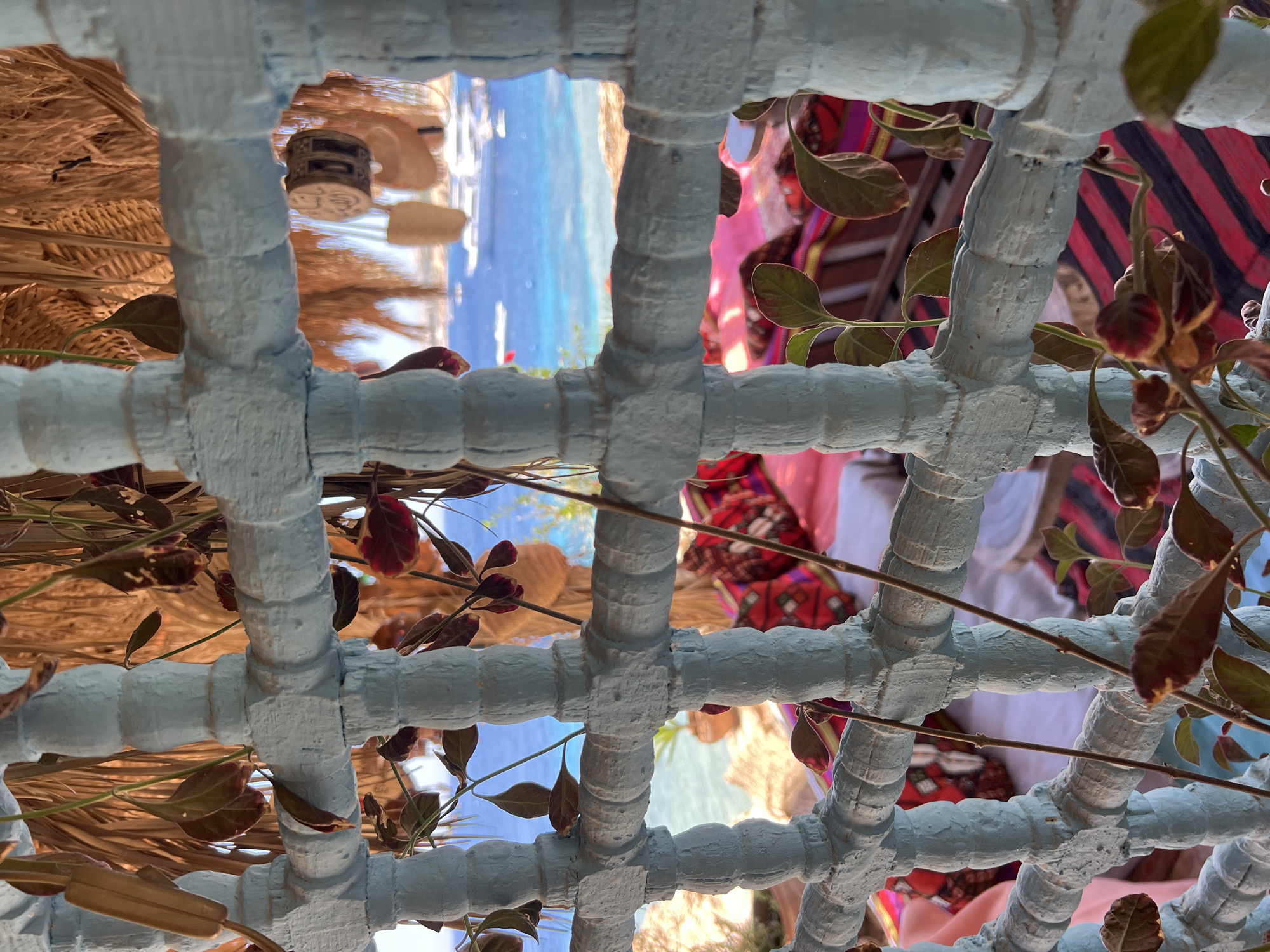 An eye-level, medium shot captures a view through a rustic, light blue, wooden lattice structure, possibly a balcony railing or screen, at an outdoor seating area in Sharm El Sheikh, Egypt. The lattice is weathered and textured, with some of the paint chipped. Several thin, dry branches with dark leaves hang in front of and through the lattice, adding a natural, somewhat overgrown feel to the scene.
Beyond the lattice, the image opens up to a vibrant seascape. The water is a brilliant turquoise and blue, stretching out to the horizon under a bright, sunny sky. Several boats, including a large white yacht, are visible on the water, indicating a popular tourist or leisure destination. In the foreground, beneath the lattice and on a lower level, there appears to be a seating area with cushions and textiles. These textiles display a mix of colors and patterns, with prominent stripes of red, black, and possibly pink, along with some richly patterned cushions that suggest a bohemian or traditional aesthetic. A thatched roof or canopy is visible above, providing shade. The overall impression is one of a relaxed, scenic spot by the sea, likely a café or restaurant, in a resort town like Sharm El Sheikh. There are no people directly visible in the frame, but the setup implies a space for human use and enjoyment.