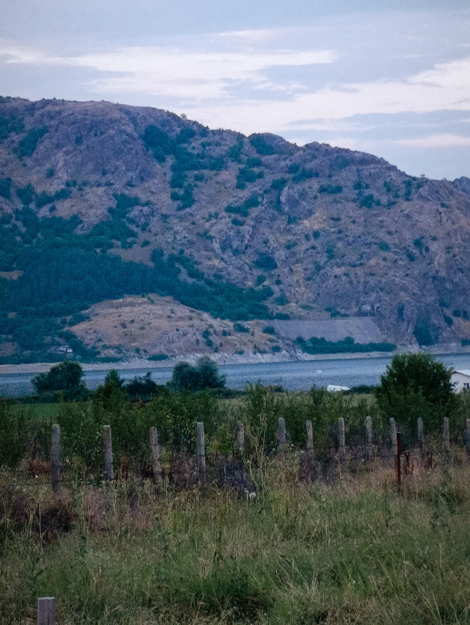 This is a landscape photograph taken in the vicinity of Shiroko pole, Bulgaria. The image depicts a large, rocky mountain covered in sparse vegetation, with a body of water in the foreground. A line of wooden fence posts and tall grass stretches across the bottom half of the frame. The sky is overcast with some visible clouds. The overall scene suggests a natural, undeveloped environment.