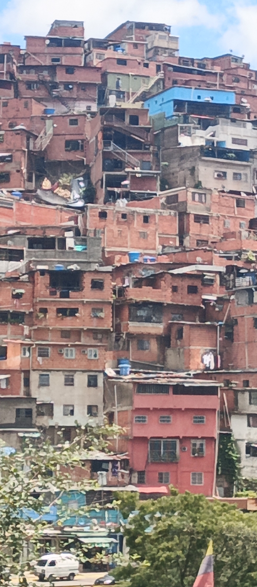 This is a medium-distance, eye-level shot of a densely packed hillside favela in Caracas, Venezuela, under a bright sky with scattered clouds. The buildings, predominantly constructed from red brick and concrete, are stacked haphazardly on the steep slope, creating a visually overwhelming and irregular urban landscape. Many of the structures appear to be multi-story, with some featuring corrugated metal or sheet metal roofing. Various shades of paint, including blue, pink, and green, are visible on some of the buildings, offering pops of color against the dominant red brick. Laundry is visible hanging from some of the structures, suggesting ongoing domestic activity. Narrow pathways and staircases weave between the buildings, indicating the challenging terrain and the informal nature of the settlement. In the foreground, lush green trees partially obscure the view of the lower sections of the favela. Below the trees, a white van is parked on a street, with another dark vehicle partially visible. The overall impression is one of vibrant, albeit precarious, urban life in a challenging environment.