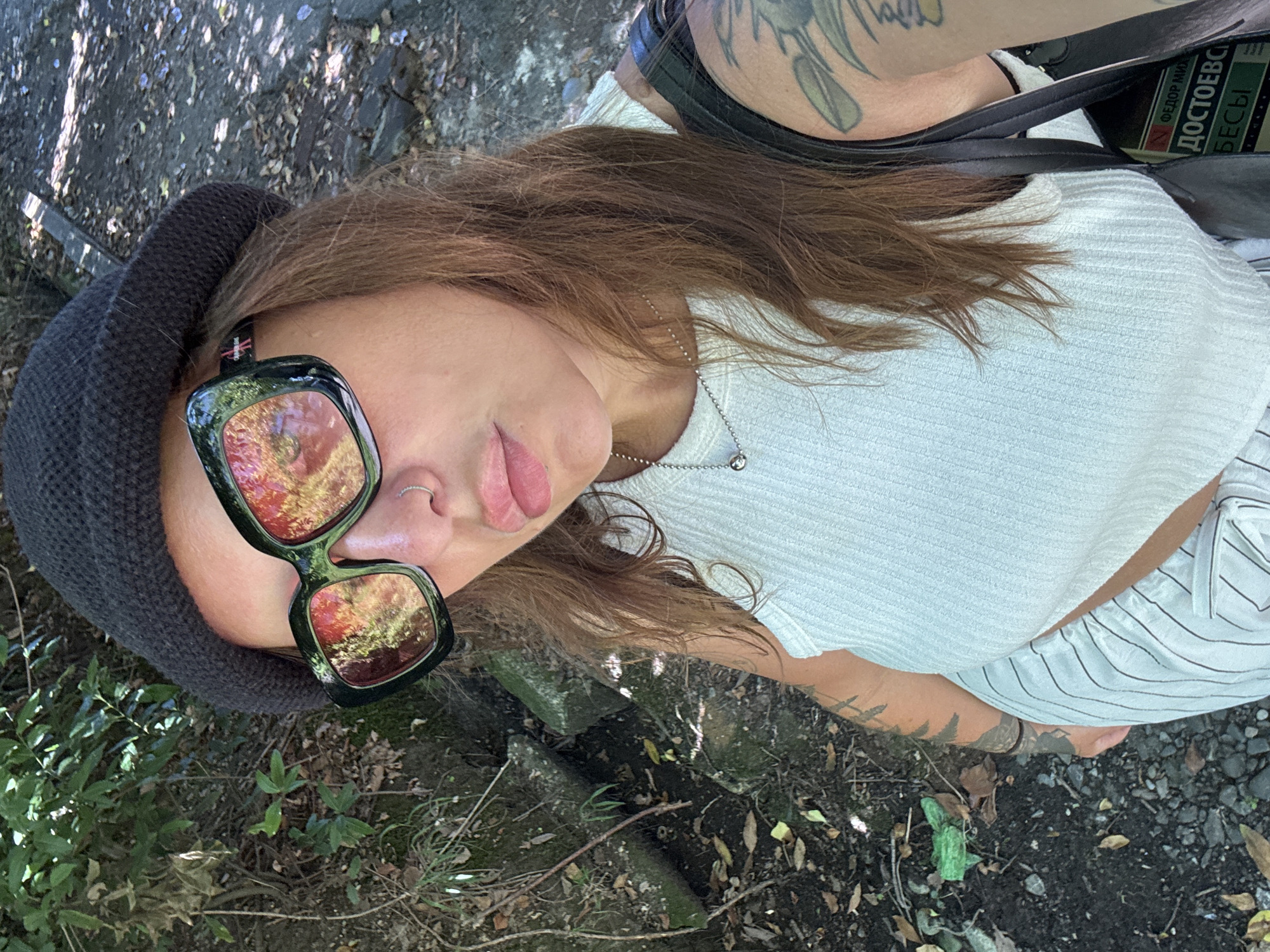A woman with brown hair, wearing sunglasses, a black beanie, and a white top, takes a selfie outdoors. She has a nose ring and is wearing a necklace. Her left arm, visible in the frame, is tattooed. She is wearing striped pants. In the background, there is foliage and a path or ground covered in small stones. Part of a black bag and a book with Russian text are visible near her right shoulder. The environment appears to be a park or natural area. The text on the book, "ФЕДОР МИХ ДОСТОЕВСИ БЕСЫ" (Fyodor Dostoevsky, "Demons" or "The Possessed"), suggests a cultural context potentially related to the city of Sochi, Russia, which has literary and cultural significance. The sunlight indicates it is daytime.