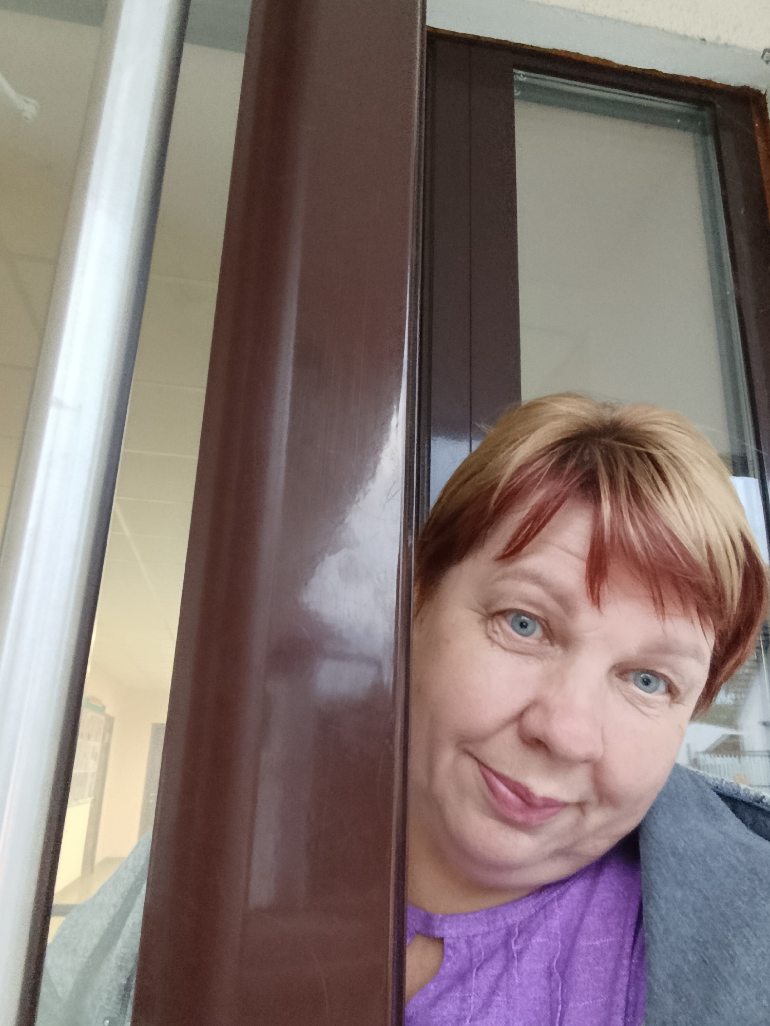 A woman with short, reddish-blonde hair and blue eyes is peeking from behind a brown door frame. She is wearing a purple top and a grey jacket. The background appears to be an interior hallway or room, with white walls and ceiling visible through a glass panel. The image is a selfie taken from a low angle. The location is identified as Svietlahorsk, Belarus.