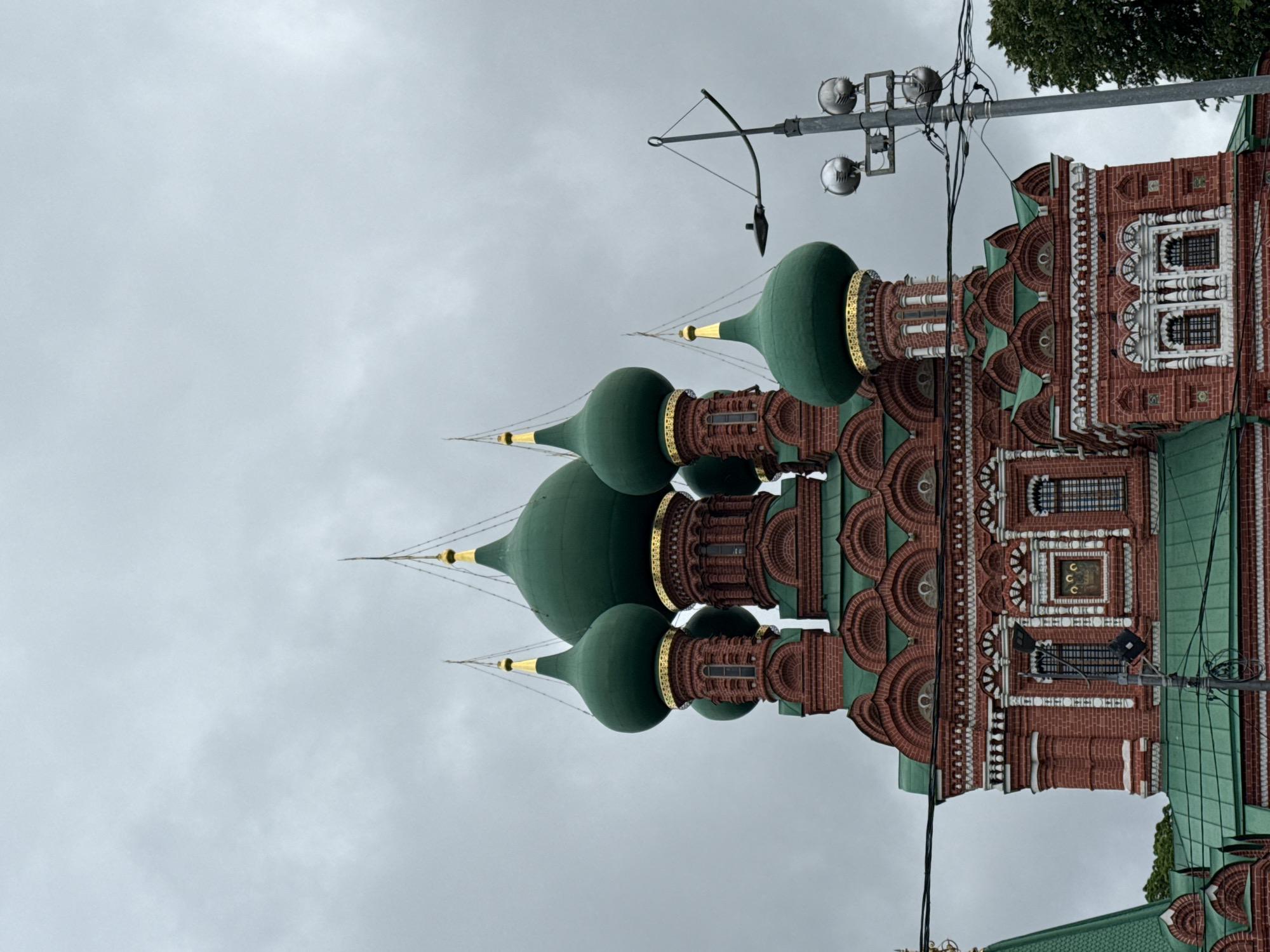 The image shows the Church of the Smolensk Icon of the Mother of God in Khimki, Russia. The architecture features multiple green onion domes adorned with gold accents. The church building is constructed of red brick, embellished with white and green architectural details. A street light pole with multiple lamps is situated near the church. The sky is overcast and gray.