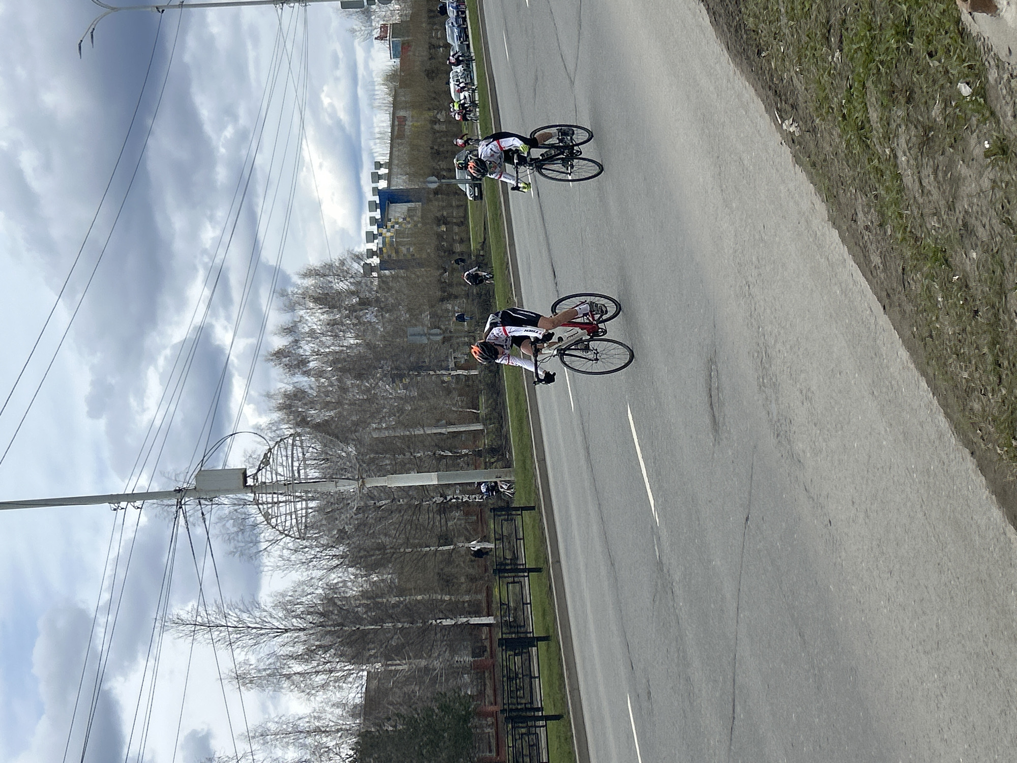 The image captures a scene in Izhevsk, Russia, featuring two cyclists riding on a paved road. The cyclists wear helmets and athletic attire. A grassy verge and a black metal fence separate the road from a line of bare trees. In the background, buildings and parked cars are visible. Overhead, utility poles and power lines stretch across a cloudy sky. The road is marked with a painted white line.