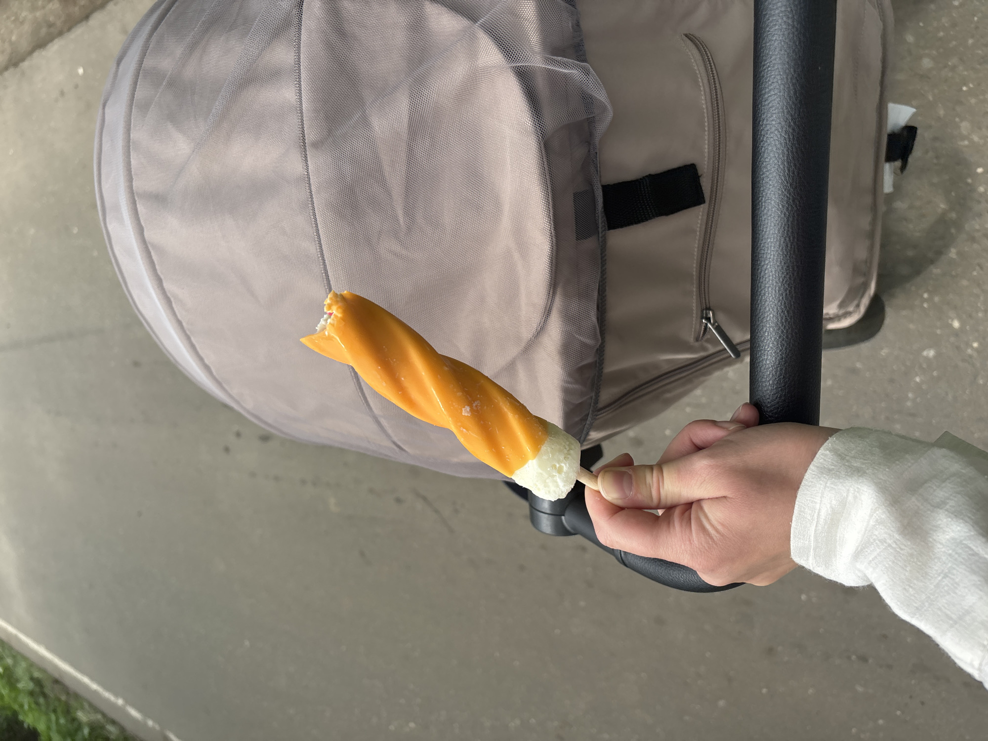 A hand in a white shirt sleeve holds an orange and white popsicle in front of a beige baby stroller with a gray mesh cover. The background appears to be a paved surface, possibly a sidewalk.