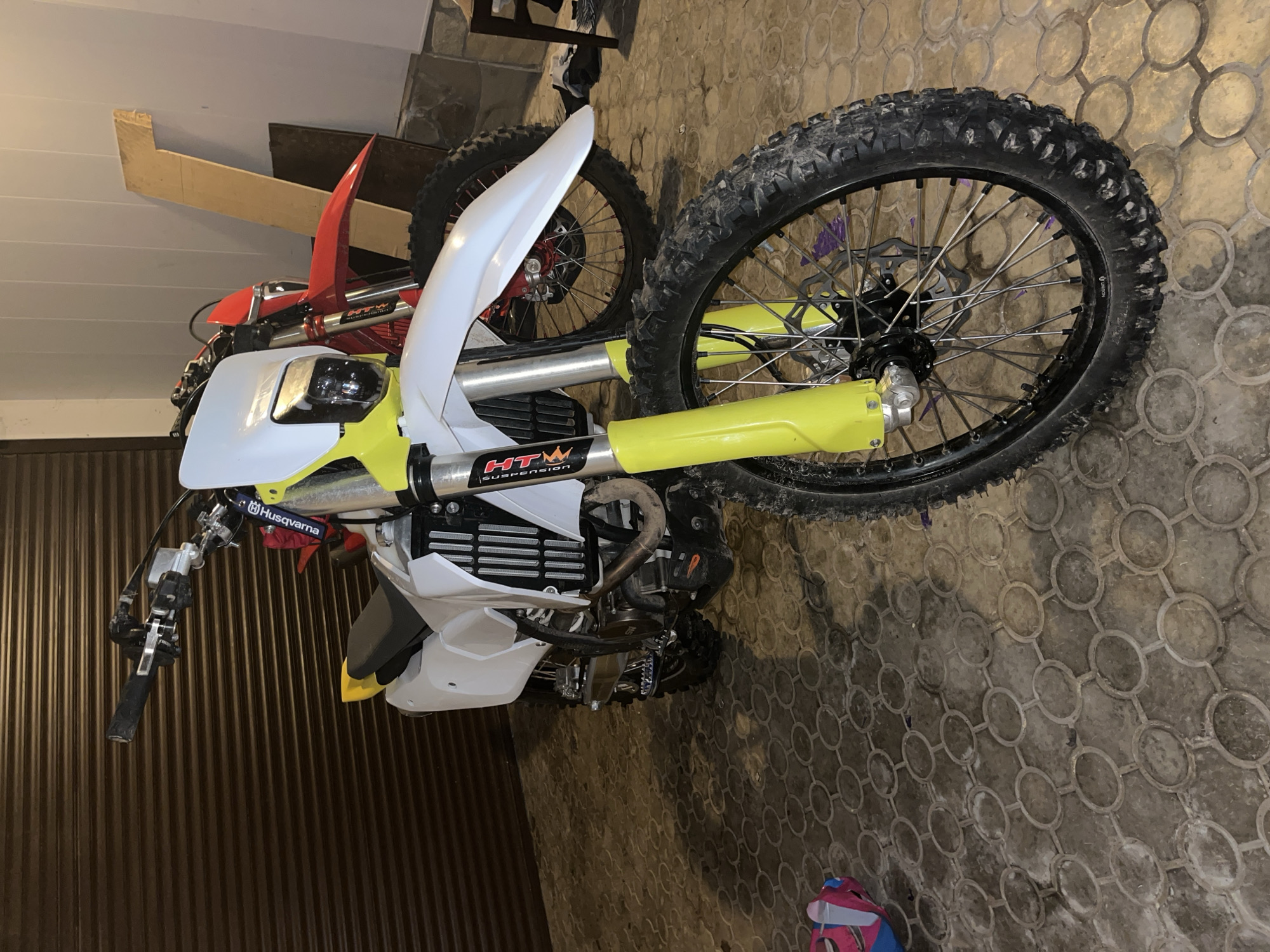 The image captures two dirt bikes parked indoors, likely in a garage or workshop in Ореховка, Russia. The primary subject is a white and yellow dirt bike, positioned diagonally across the frame. Its front white fender and headlight are visible, along with bright yellow front suspension forks. Black knobby tires indicate its off-road purpose. On the handlebars, a blue strap or tag displays "Husqvarna," and on the front fork, a sticker reads "HTW SUSPENSION" with a crown logo. The bike's engine area, including a black radiator grille and exhaust pipe, is visible beneath the white fairing.
Behind the prominent bike, parts of a second dirt bike can be seen, notably a red front fender and a wheel with a red hub.
The floor of the setting is a textured pavement with an interlocking circular pattern, appearing somewhat worn or dusty. On the left side, a dark, corrugated wall, possibly a garage door or metal siding, occupies the background. Above this, a lighter wall and some wooden structures are discernible. In the bottom right corner, a small, colorful object, possibly a shoe or a piece of clothing, lies on the floor. The bikes are stationary, indicating they are parked or stored, rather than being actively ridden. The lighting suggests an indoor, artificial environment.