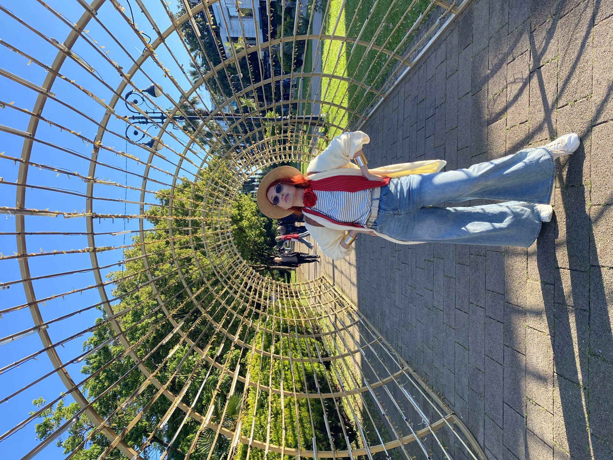 This image captures a bright, sunny outdoor scene in Sochi, Russia, featuring a woman posing within a decorative archway. The primary subject is a woman standing on a paved walkway on the right side of the frame, looking towards the camera. She has reddish hair, is wearing sunglasses, and a straw hat with a dark band. Her attire includes a striped top, wide-leg light blue denim jeans, and white sneakers. A light-colored, possibly cream, cardigan is draped over her shoulders, and a vibrant red scarf or sash with a red rose embellishment is visible on her chest. A gold-toned cuff bracelet adorns her right wrist.
The setting is dominated by a large, semi-circular archway or tunnel structure made of light-colored gridwork, possibly metal or bamboo, stretching into the background. This structure is adorned with numerous small, yellow-orange lights. Lush green trees and foliage are visible through and on either side of the archway, with a clear blue sky overhead. A street lamp with two fixtures is visible through the gridwork against the sky. The paved walkway beneath the archway is made of dark grey interlocking pavers, with distinct shadows of the gridwork cast upon it, indicating bright sunshine. In the background, several blurred figures suggest other people are present further along the walkway.
The activity depicted is the woman posing for a photograph in a scenic, decorative environment. The time of day is clearly sunny daytime, likely mid-day or afternoon, given the bright light and strong shadows. The weather is clear and pleasant. No visible text is present in the image.