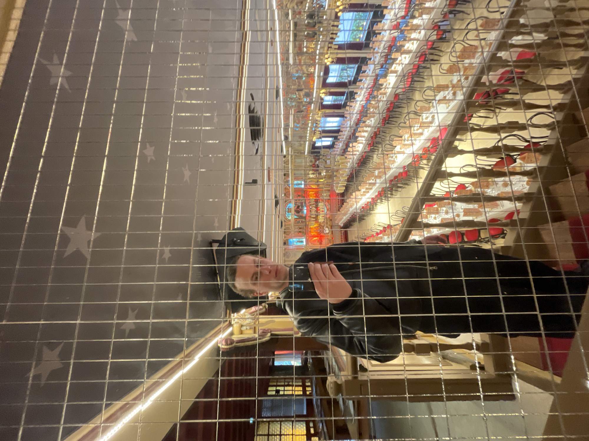A person in a black jacket and cap stands behind a grid of mirrored squares. The reflection in the mirrors shows a grand interior with rows of what appear to be desserts, possibly pastries or candies, displayed on shelves or racks. The reflection extends up, revealing architectural details like windows and ornate fixtures. The walls and ceiling in the reflection suggest a luxurious or historic establishment. Some of the squares have star designs on them. Based on the image content, there is no proof that this scene is taking place in Collonges-au-Mont-d'Or, France.