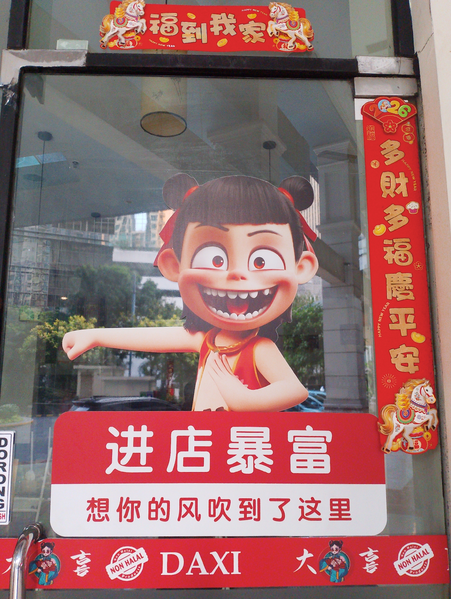 This is a medium shot of a storefront with festive Chinese New Year decorations. The primary focus is a large, cut-out image of a cartoon girl with dark hair in pigtails and a wide, toothy grin. She's wearing a red outfit and has her arm extended forward.
The decorations are predominantly red and gold. A banner above the cartoon reads "福到我家" (Fortune Comes to My Home) with a golden horse decoration. To the right, a vertical banner reads "多财多福, 膺平平安安" (Abundant wealth and blessings, smooth and peaceful throughout the year). This banner also features smaller decorative elements like coins, a cupcake, and a horse.
Below the cartoon is a large red sign with white text that reads "进店暴富" (Enter the store for great wealth) and "想你的风吹到了这里" (The wind of thinking of you has blown here).
At the bottom, there are smaller red banners. One features a cartoon character and Chinese characters. Another banner has the text "DAXI" and a "NON HALAL PRODUCTS" seal.
The storefront appears to be a glass door or window, reflecting the environment outside, which includes trees and buildings. The overall scene suggests a business celebrating Chinese New Year. The presence of "NON HALAL PRODUCTS" suggests this may be a non-Muslim owned business in an area with a significant Muslim population, or a business that caters to a diverse clientele. Based on the typical business names and decorations found in South Jakarta, Indonesia, this is likely a shop or restaurant in that area.
