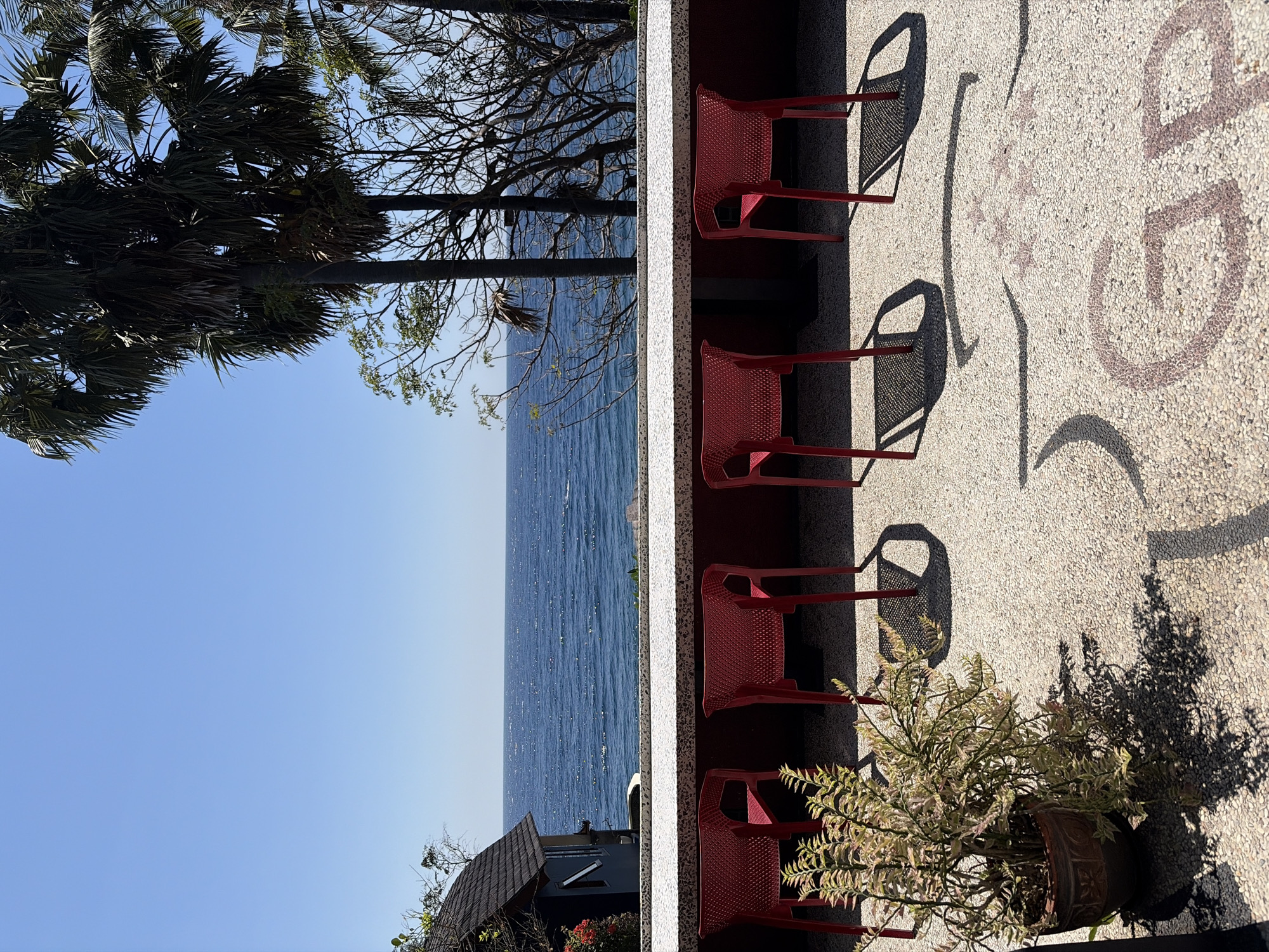 A sunny, outdoor scene in Abang, Indonesia, shows a row of five red plastic chairs facing a stone counter with a view of the ocean. The chairs are evenly spaced and cast distinct shadows on the pebble-textured ground. To the left of the chairs, a potted plant with variegated leaves sits near the counter. Behind the counter, the blue ocean stretches to the horizon under a clear sky. Palm trees and other vegetation frame the view of the ocean. A dark-colored building with a dark roof is partially visible to the far left, behind the plant and in front of the ocean. The ground has a pattern embedded within the pebbles, with parts of letters visible, possibly forming a word or logo.