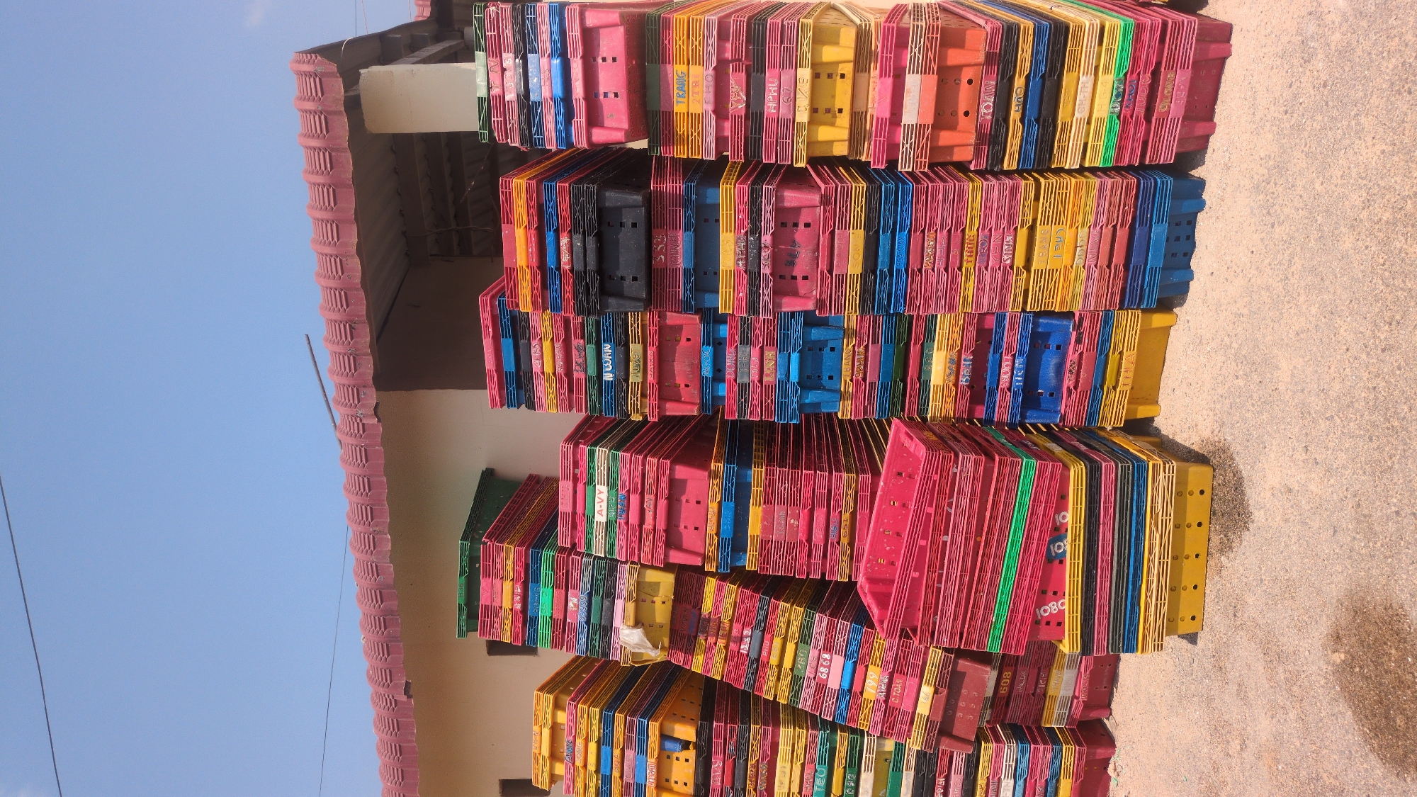 The image depicts stacks of colorful plastic crates in what appears to be Phước Dinh, Vietnam, given the prompt's reference to this city. The crates, primarily red, yellow, blue, and green, are stacked unevenly, reaching towards the blue sky. They appear to be next to a building with a red-tiled roof. The ground is a light-colored, possibly concrete or dirt surface. The lighting is bright, indicating daytime. No people are visible in the frame.