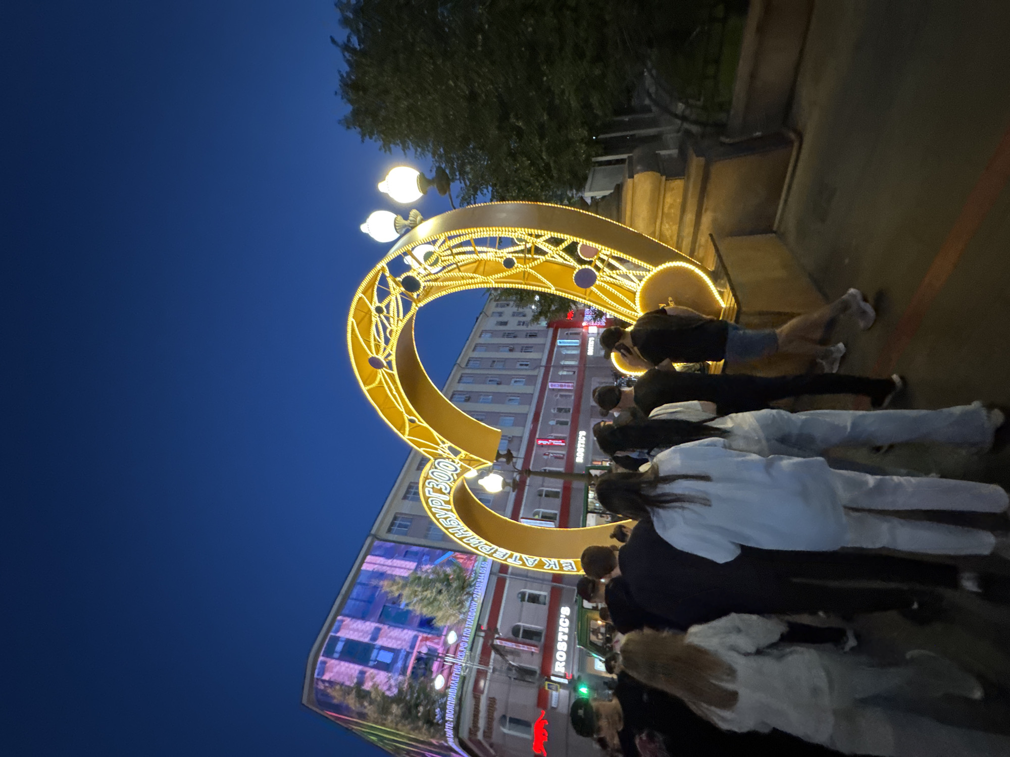 In Yekaterinburg, Russia, at night, a large illuminated heart-shaped art installation is the central focus on a city street. The structure is gold and adorned with yellow lights, intricate patterns, and circular accents. It is positioned in front of a multi-story building with a digital display projecting an image of trees and a building facade. Below this, the building features several businesses, including one labeled "ROSTIC'S". A crowd of people, mostly seen from behind, are gathered around the installation, with some looking towards it and others walking past. The sky above is a deep blue, indicating twilight or night. Streetlights and the building's lighting cast a glow on the scene.