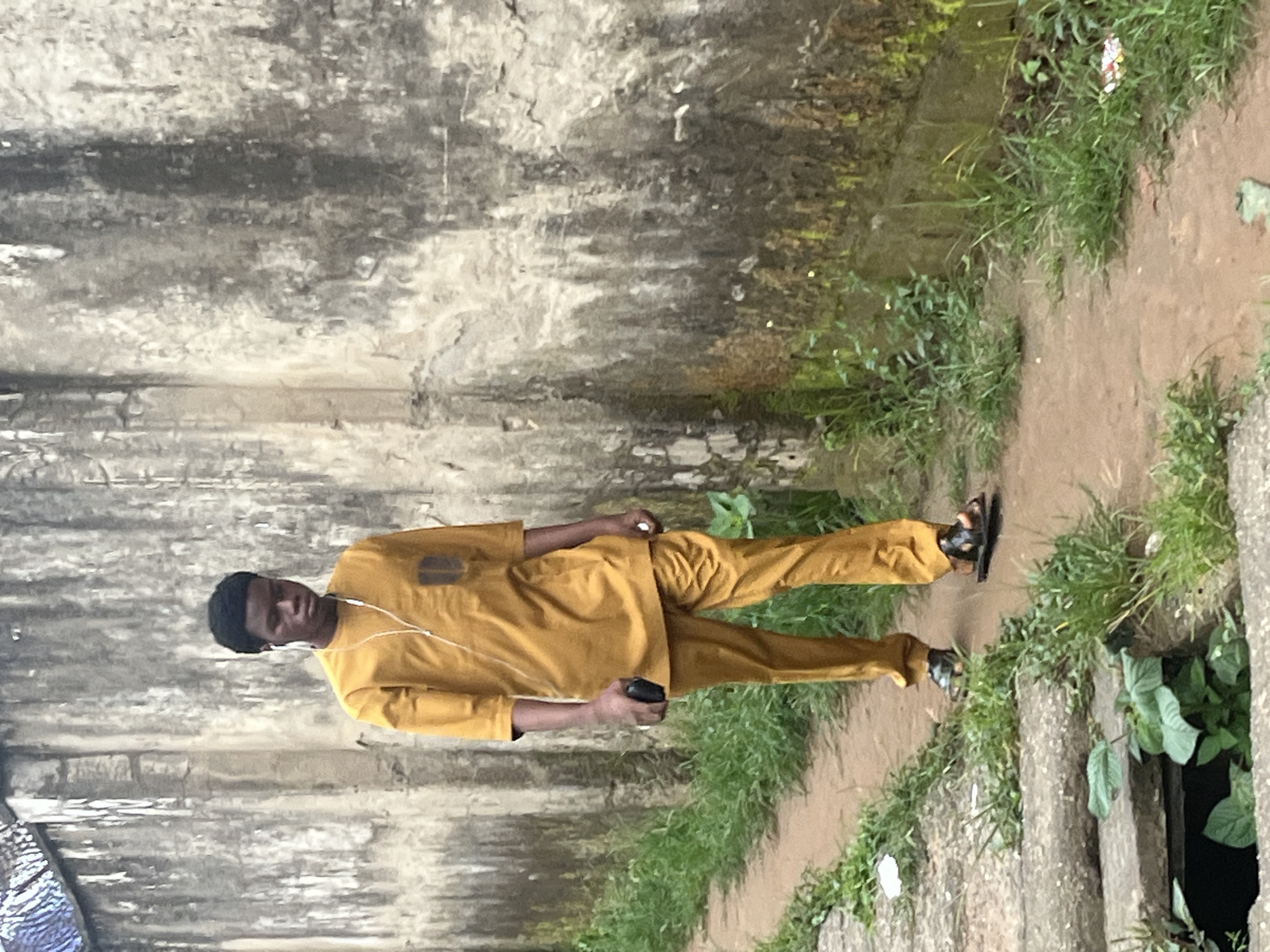 A young Black male walks along a dirt path next to a rough concrete wall. He is wearing a mustard-yellow tunic and matching trousers, with a dark brown square patch on the left breast pocket of the tunic. White earbuds are in his ears, and a white cord runs down his front. In his right hand, he holds a black rectangular object, possibly a phone. He is wearing dark sandals with a patterned sole. The path is lined with green grass and vegetation. The wall is textured and weathered, with patches of dirt and grime. The scene appears to be in an outdoor urban setting.