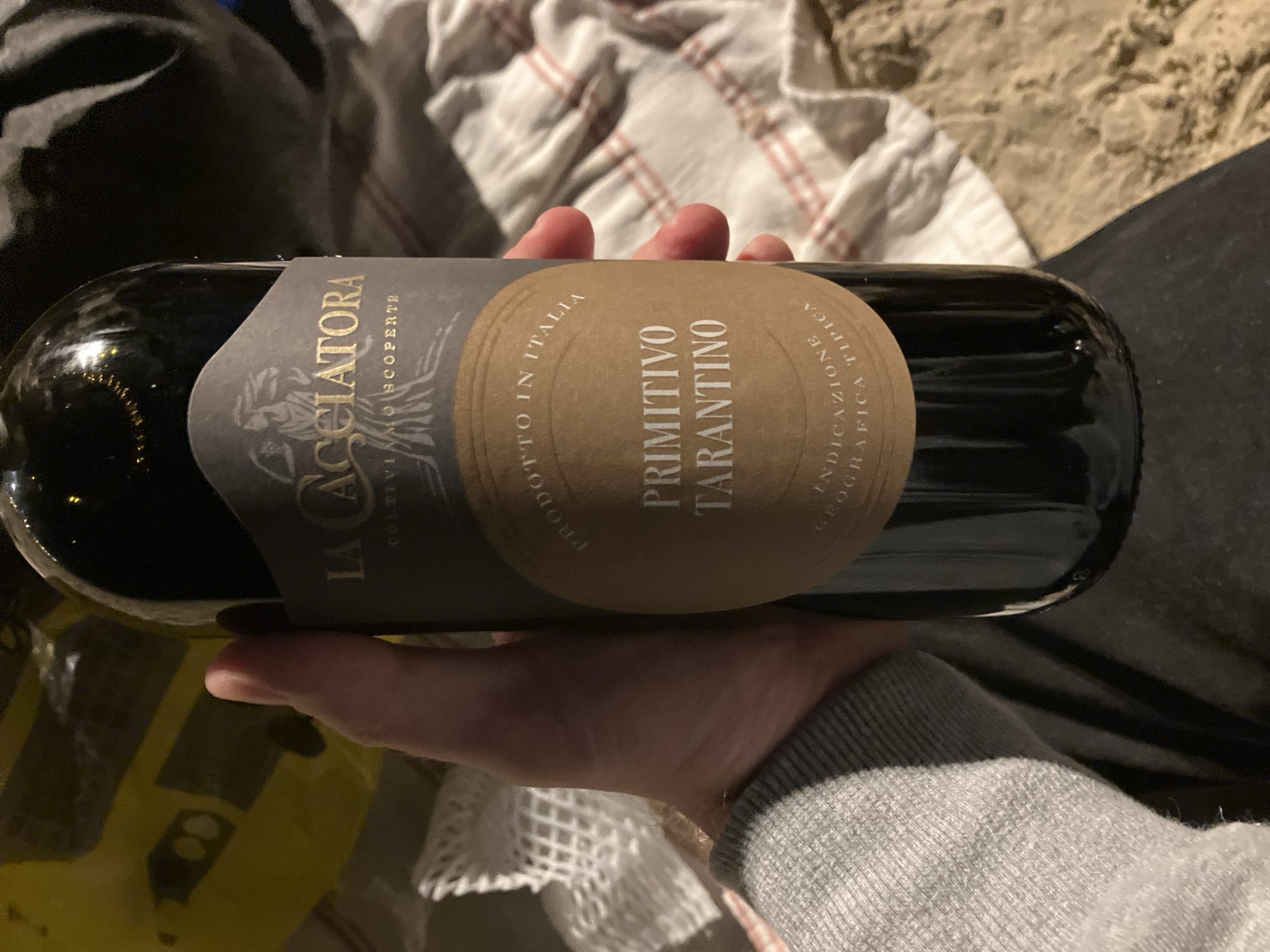 A person is holding a bottle of "La Cacciatora Primitivo Tarantino" wine. The background includes a striped white cloth and what appears to be beach sand, possibly indicating the wine is being held at a beach or outdoor setting. There is no information in the image suggesting the location is Tel-Aviv, Israel.