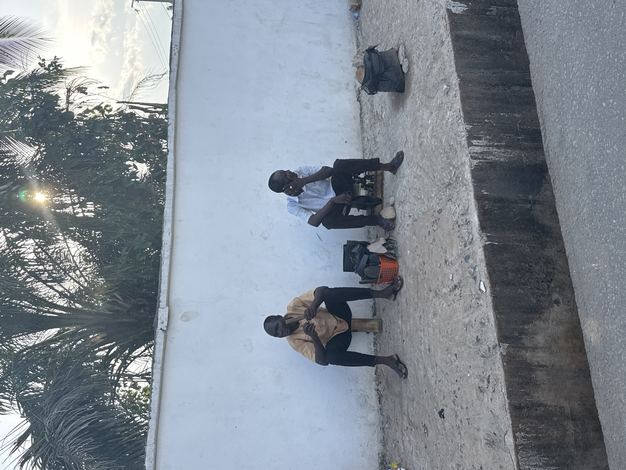 Two men are sitting on small wooden stools against a white wall. One man is wearing a light-colored shirt and dark pants and appears to be talking on a cell phone. The other man is wearing a tan shirt and dark pants. There are bags and other items behind them. Palm trees are visible on the left side of the frame. The location is Onitsha, Nigeria.
