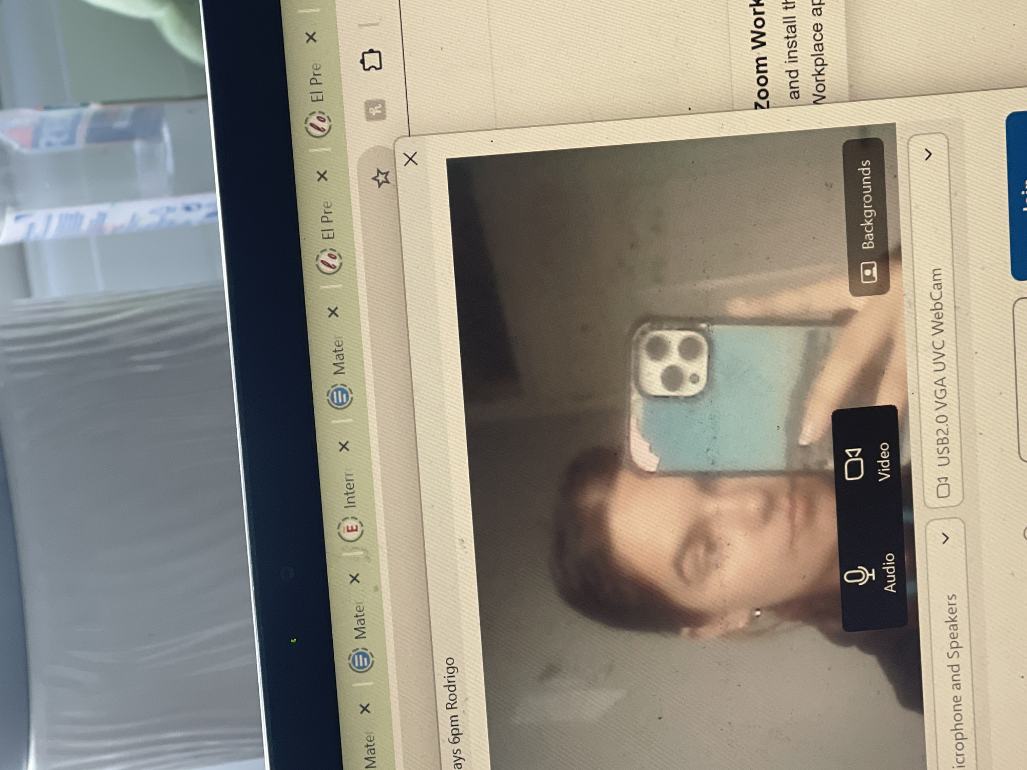 The image shows a close-up of a laptop screen. Several browser tabs are visible along the top, including those with "Mater" and "El Pre" in the title. A Zoom window is open, showing a person's face in the camera view, which is reflecting the person's phone. The Zoom interface also displays audio, video, background, and webcam options. The laptop is positioned on what appears to be a white surface. There is no information to suggest the location is in Maidstone, United Kingdom.