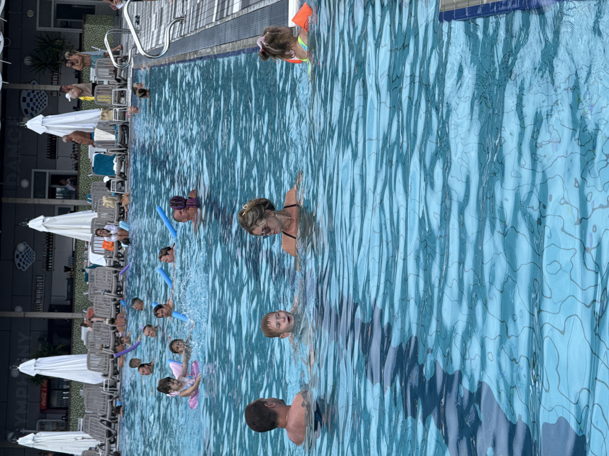 The image shows a crowded outdoor swimming pool, possibly in Массандровский поселковый совет, Ukraine, given its likely location with a bar and lounge area in the background. Several people are visible in the pool, including adults and children. Some individuals are swimming, while others are interacting with pool noodles. A young girl in a pink flotation device is in the foreground, playing in the water. A man and woman are standing behind her, also in the pool. Further back, more people are swimming and playing. In the background, there is a bar area with white umbrellas and lounge chairs. The water is clear blue, and the sun is shining.