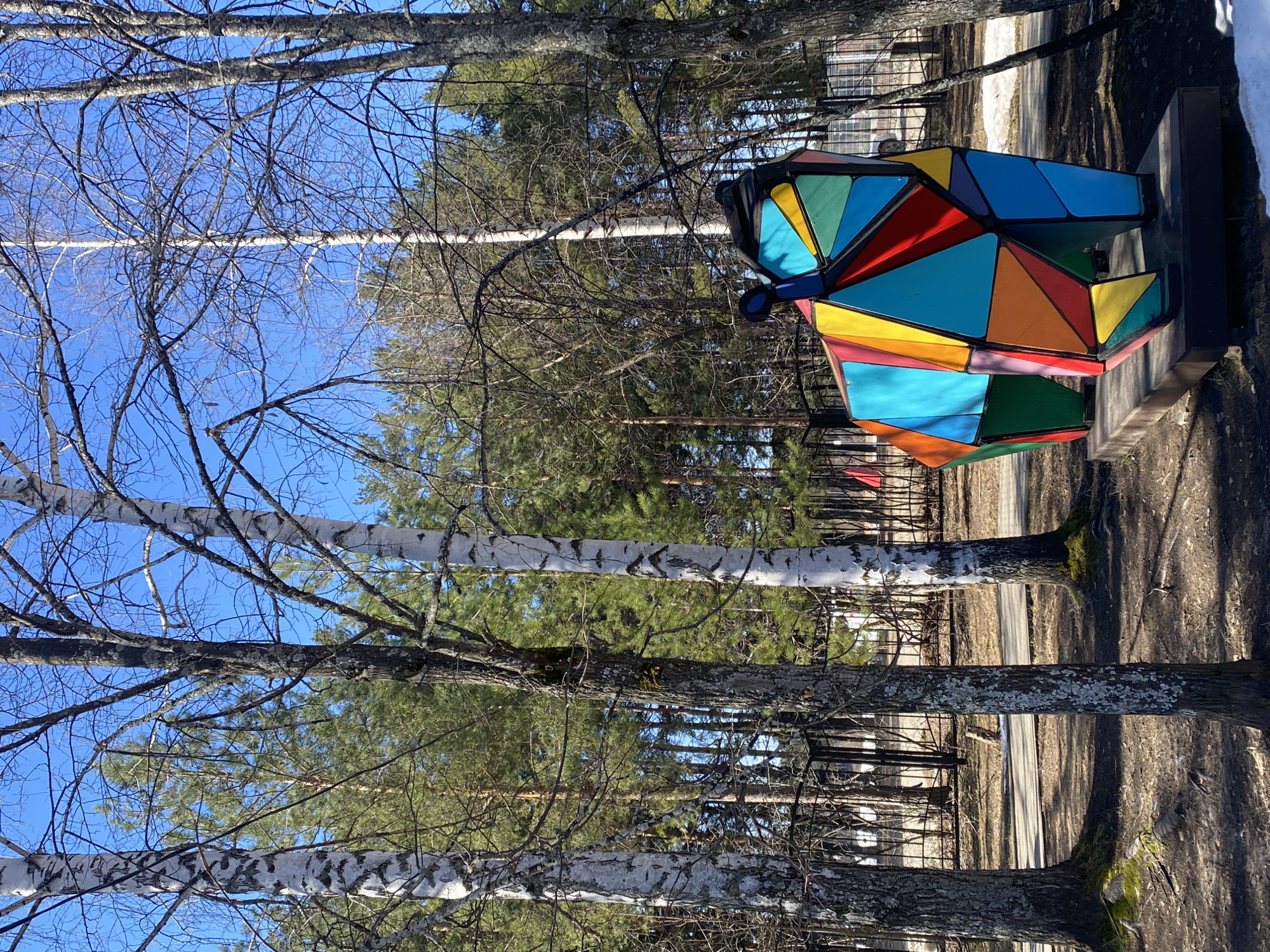 A colorful, triangular geometric sculpture sits on a concrete base in a park in Chaikovsky, Russia. The sculpture is composed of various triangular panels in blue, green, yellow, orange, and red, outlined in black. The park setting includes tall trees, mostly birch and evergreens. The sky is visible through the branches, and a path winds through the trees. A fence is visible in the background. Patches of snow are present in the upper right corner.