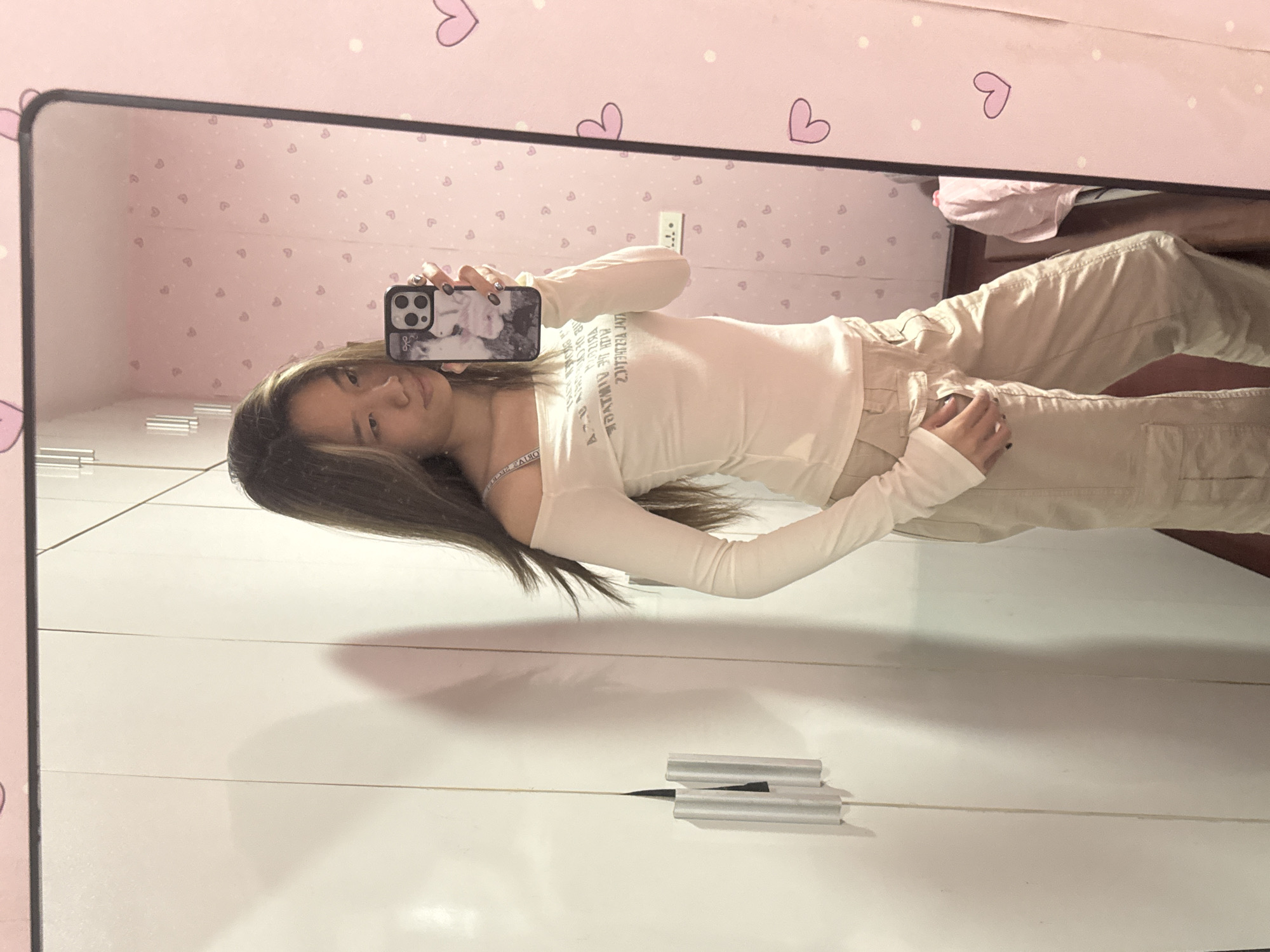 A young woman is taking a mirror selfie. The reflection shows her wearing an off-white, off-the-shoulder, long-sleeved top and tan cargo pants. She is holding a smartphone with a patterned case in her right hand. Her hair is long and brown with blonde highlights. The background includes a pink wall with a heart pattern and white cabinets. Two metallic objects that appear to be makeup are placed at the bottom, under the cabinet. The location appears to be inside a building. The city of Shapingba District, China is not discernible from the provided image.