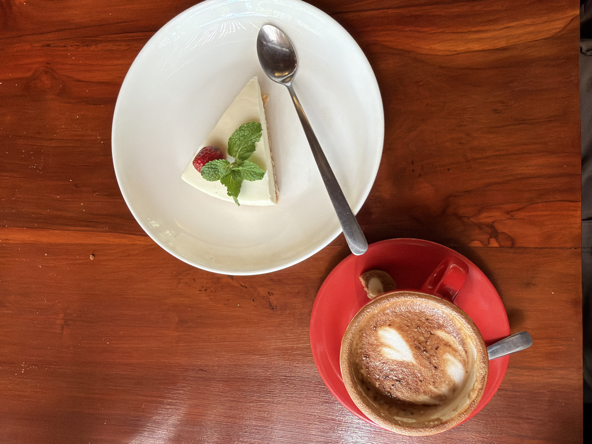 The image depicts a flat lay of a cheesecake slice on a white plate with a spoon and a cappuccino in a red cup on a matching saucer, set on a wooden table. The cheesecake is topped with a strawberry and mint leaves. The cappuccino has a heart-shaped foam design. The scene appears to be set at a café or similar establishment, but there is no further information suggesting its location in Kuta Selatan, Indonesia. There are no people visible in the image.